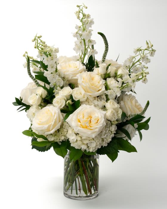Tall arrangement of white and cream roses, hydrangeas, and delphinium in a clear glass vase