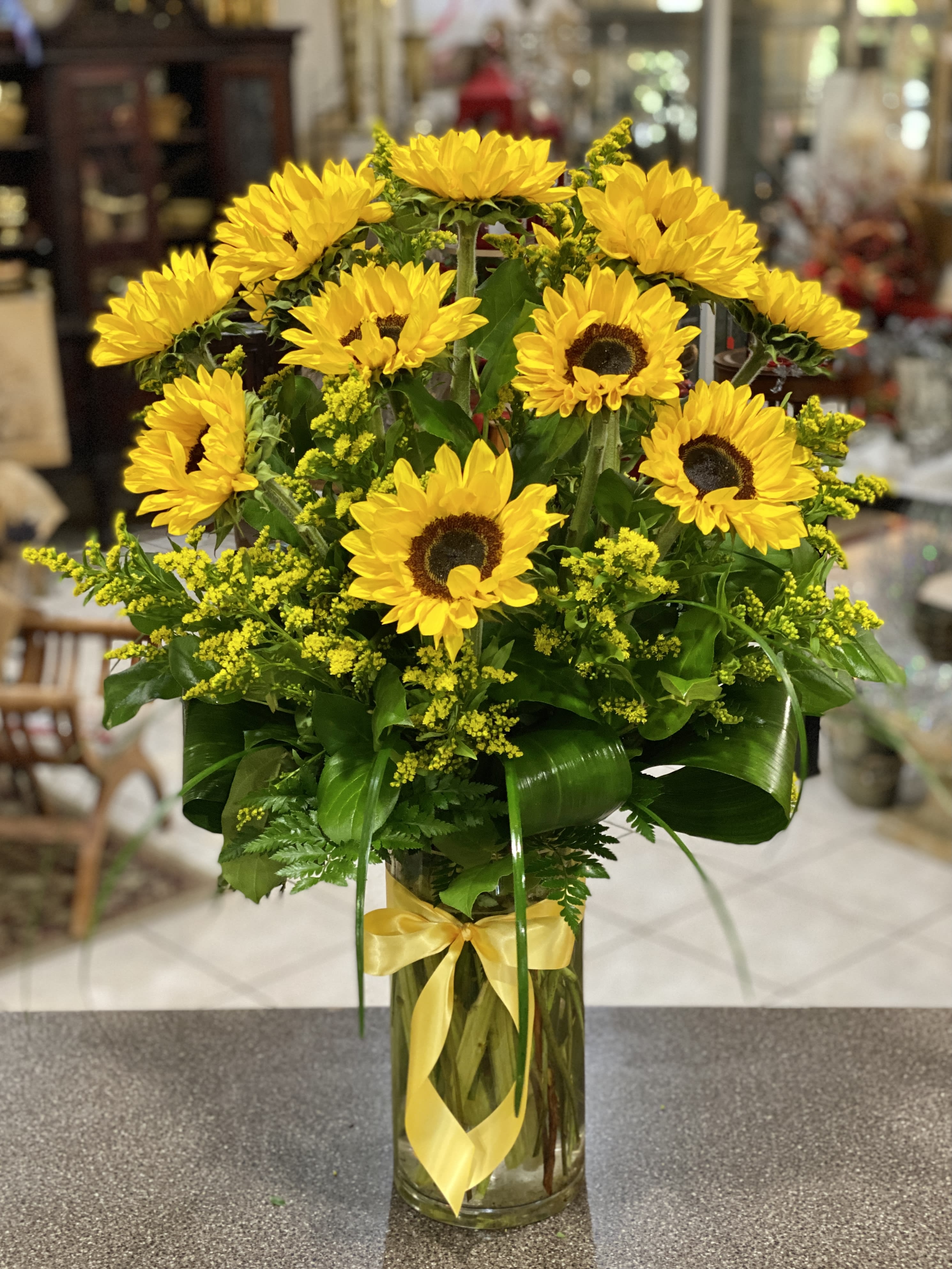 Tall arrangement of yellow sunflowers with yellow filler in a clear glass vase tied with a yellow ribbon.
