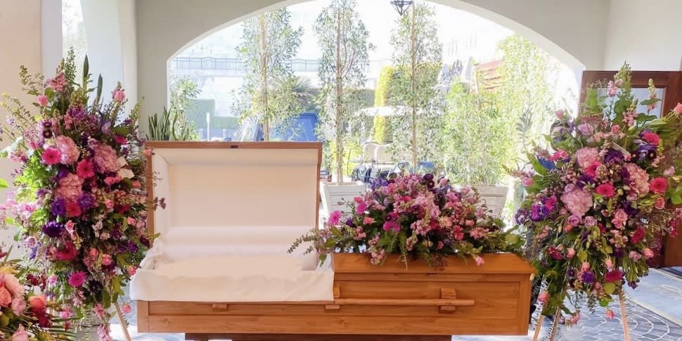 Floral casket spray with pink, purple, and white flowers beside an open casket