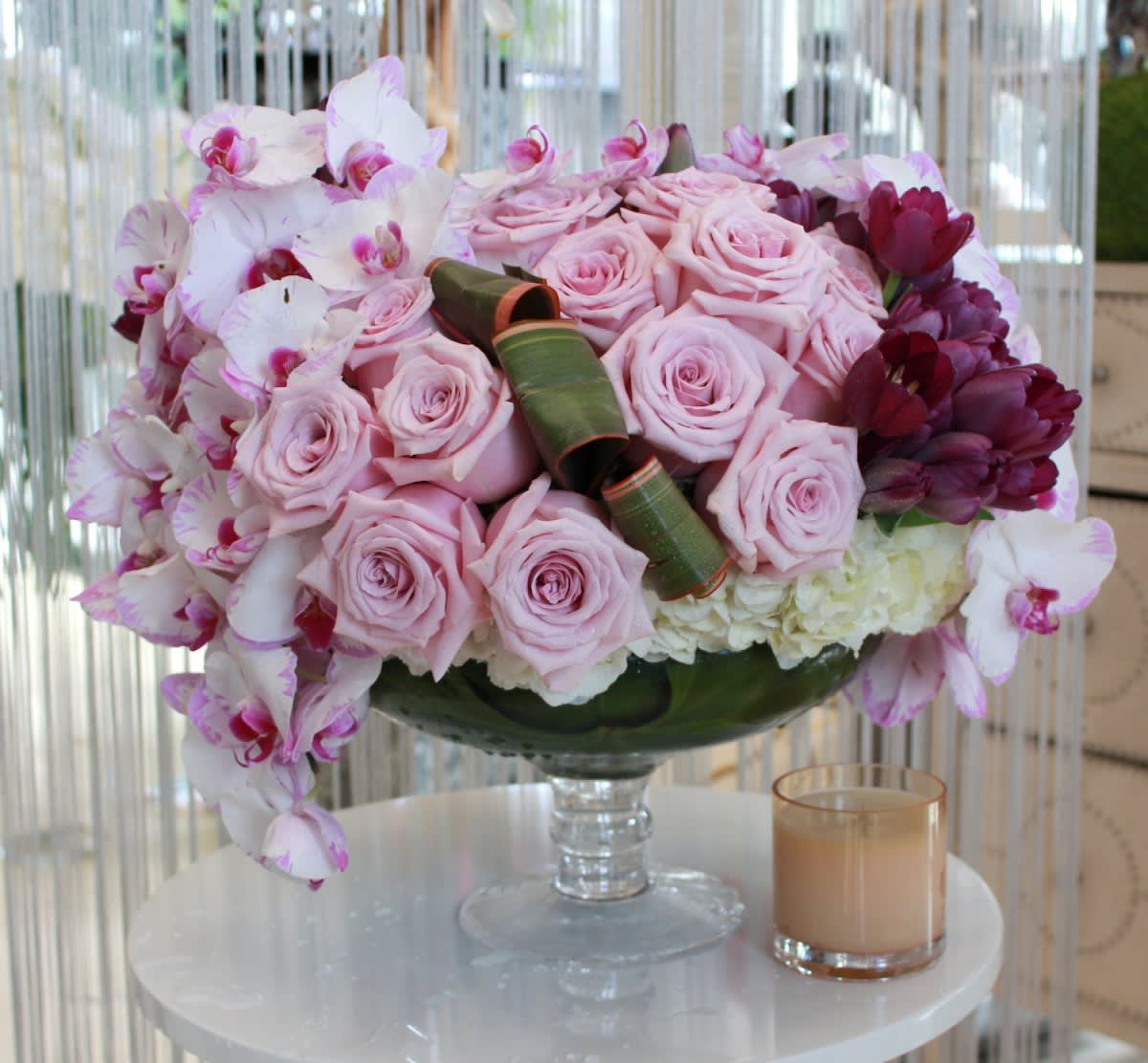 Pink roses and orchids arranged in a glass vase with a candle beside it.