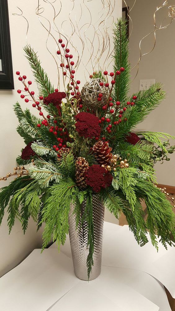 Tall holiday arrangement with red berries, pine, and pinecones in a silver vase