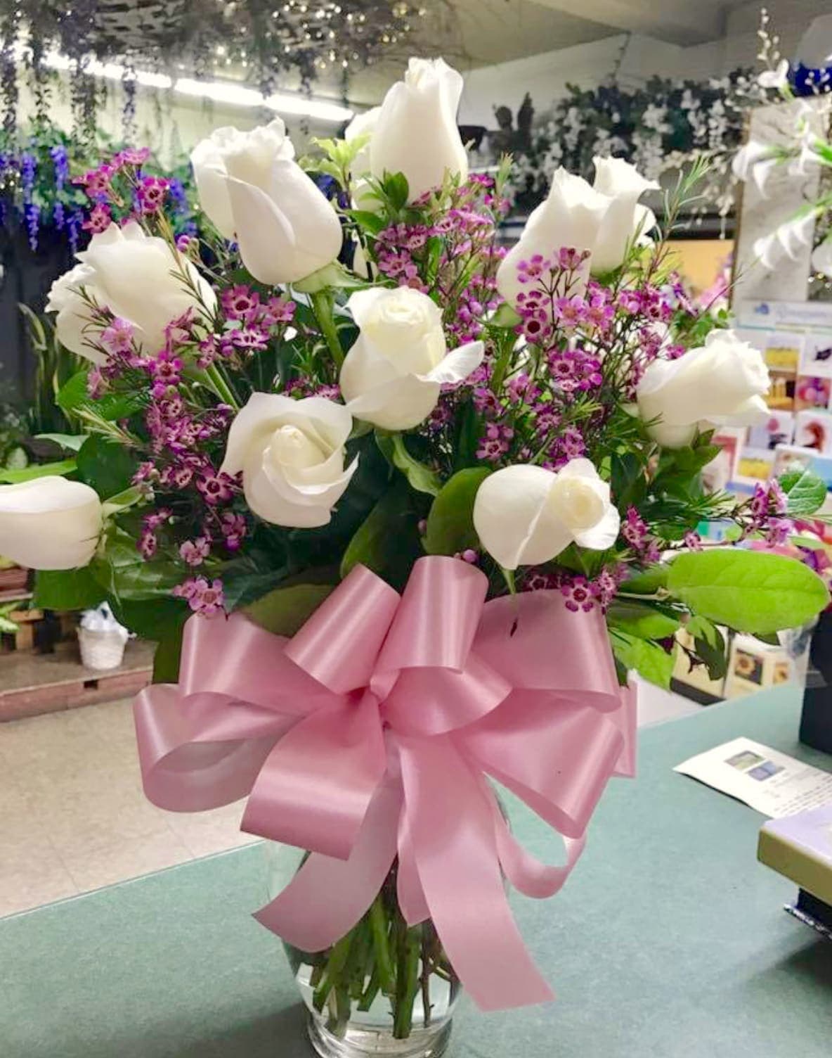 White roses and purple filler flowers in a glass vase with a pink bow