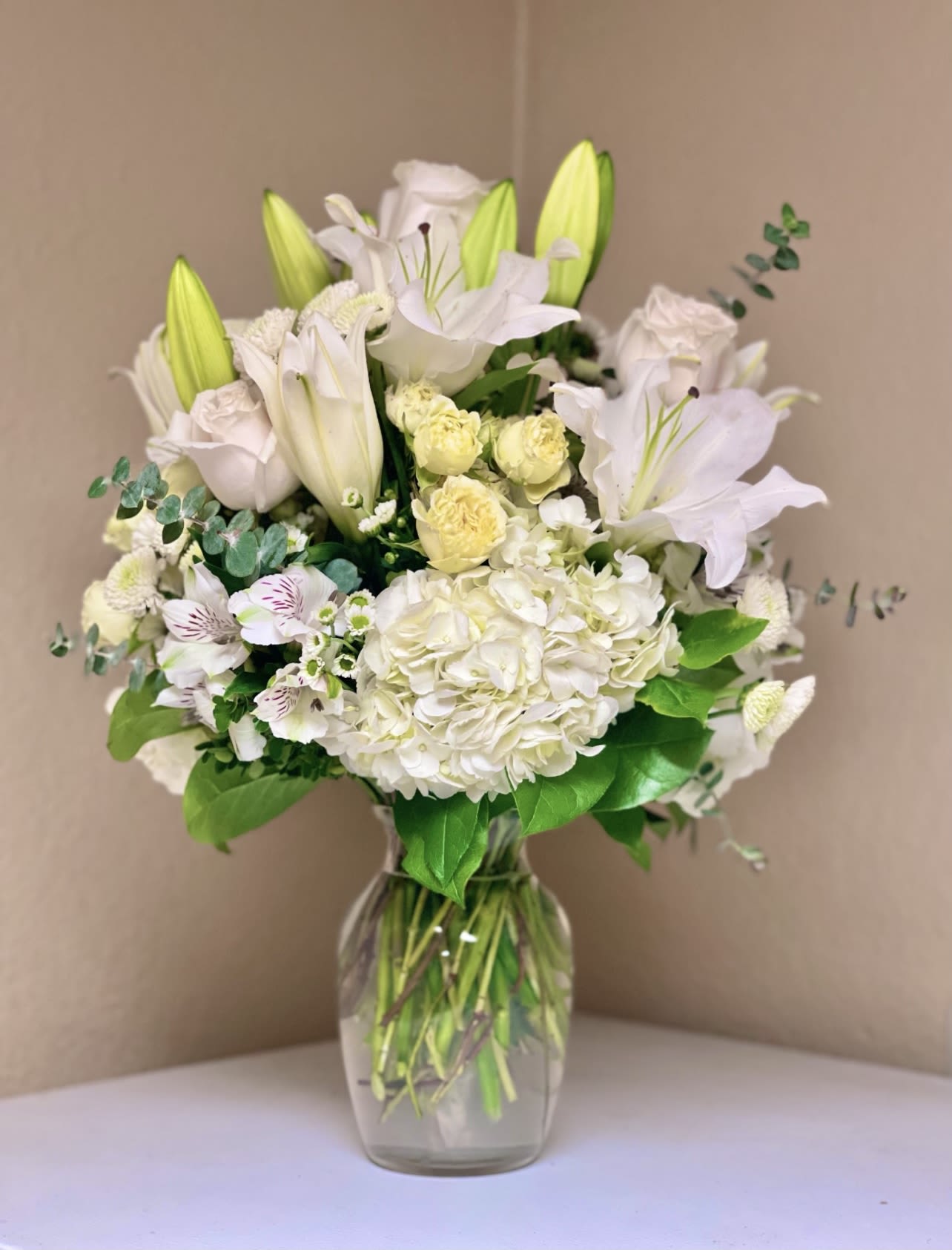 Pure Grace - There's something beautiful about an all white floral arrangement. This arrangement is filled with fresh cut Roses, spray roses, hydrangea, alstroemeria, lilies and beautiful greenery. Vase is 9.5 clear glass vase. 