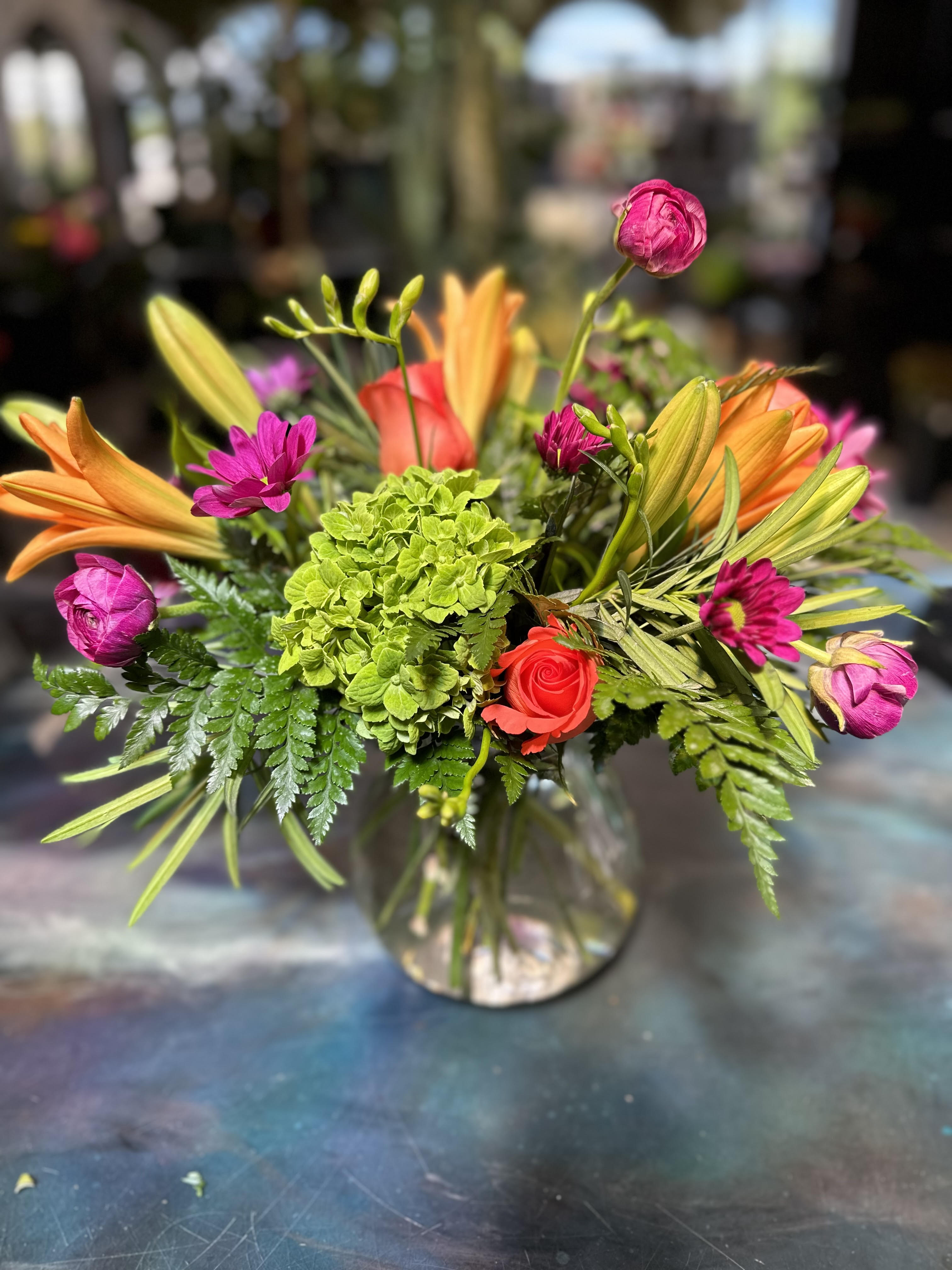 Watermelon Punch - Designed using orange asiatic lilies, green hydrangeas, coral charm roses, purpely ranunculus, and moody purple daisies, with lush, and textural foliage.