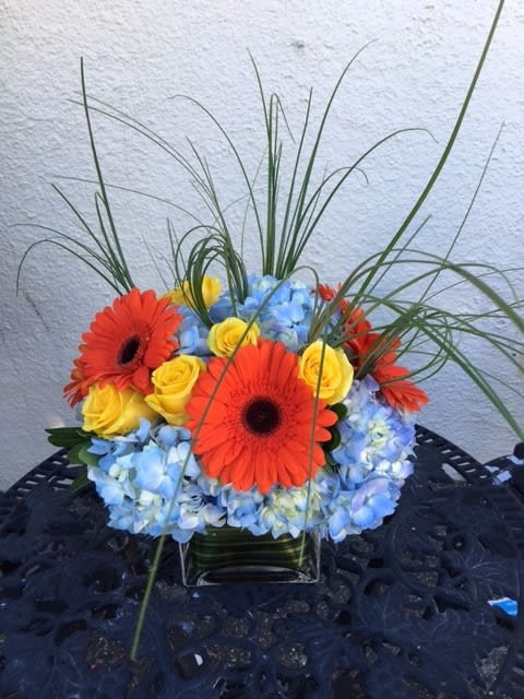 Bright bouquet of orange gerbera daisies, yellow roses, and blue hydrangeas in a glass vase