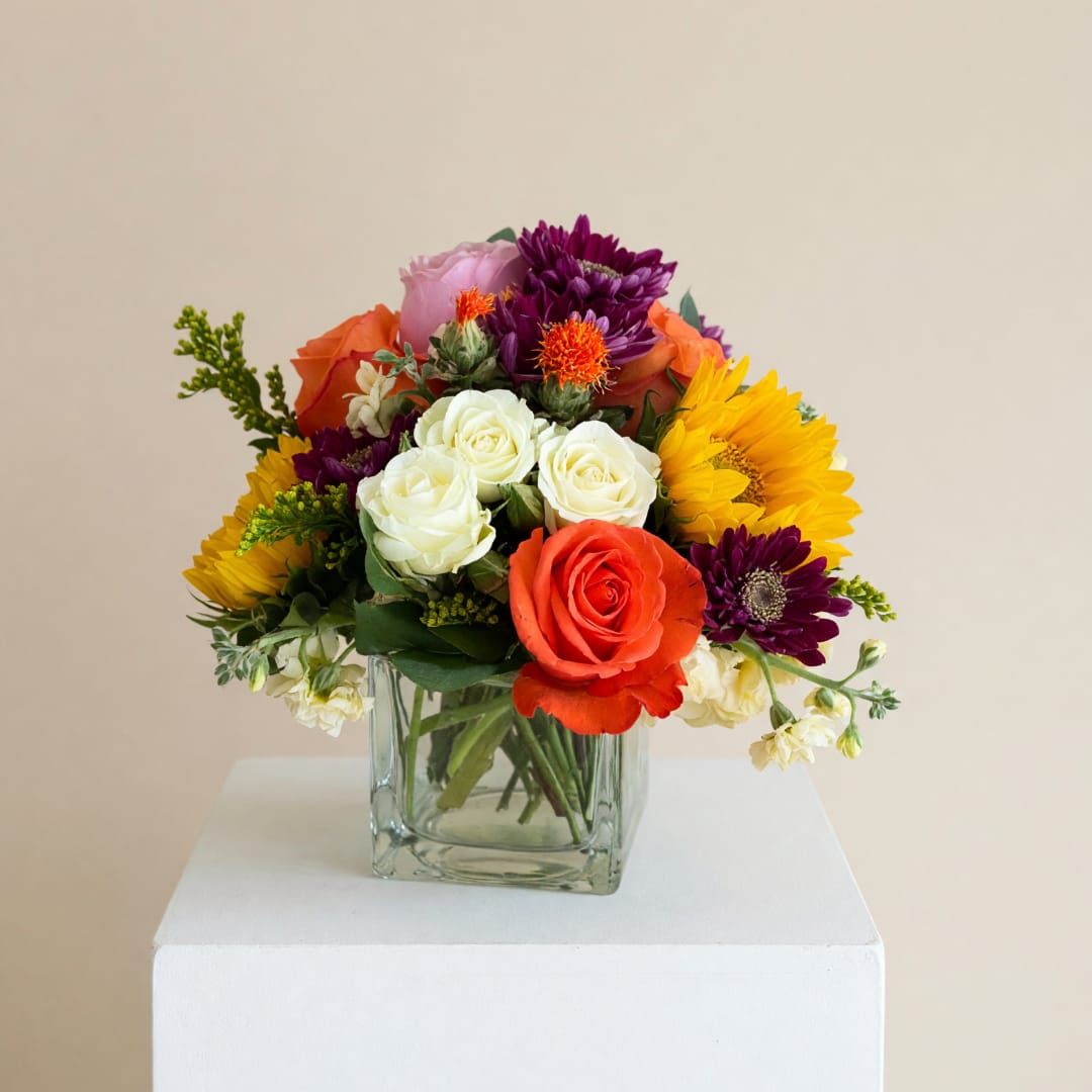 Mixed bouquet of roses, sunflowers, and purple daisies in a glass vase