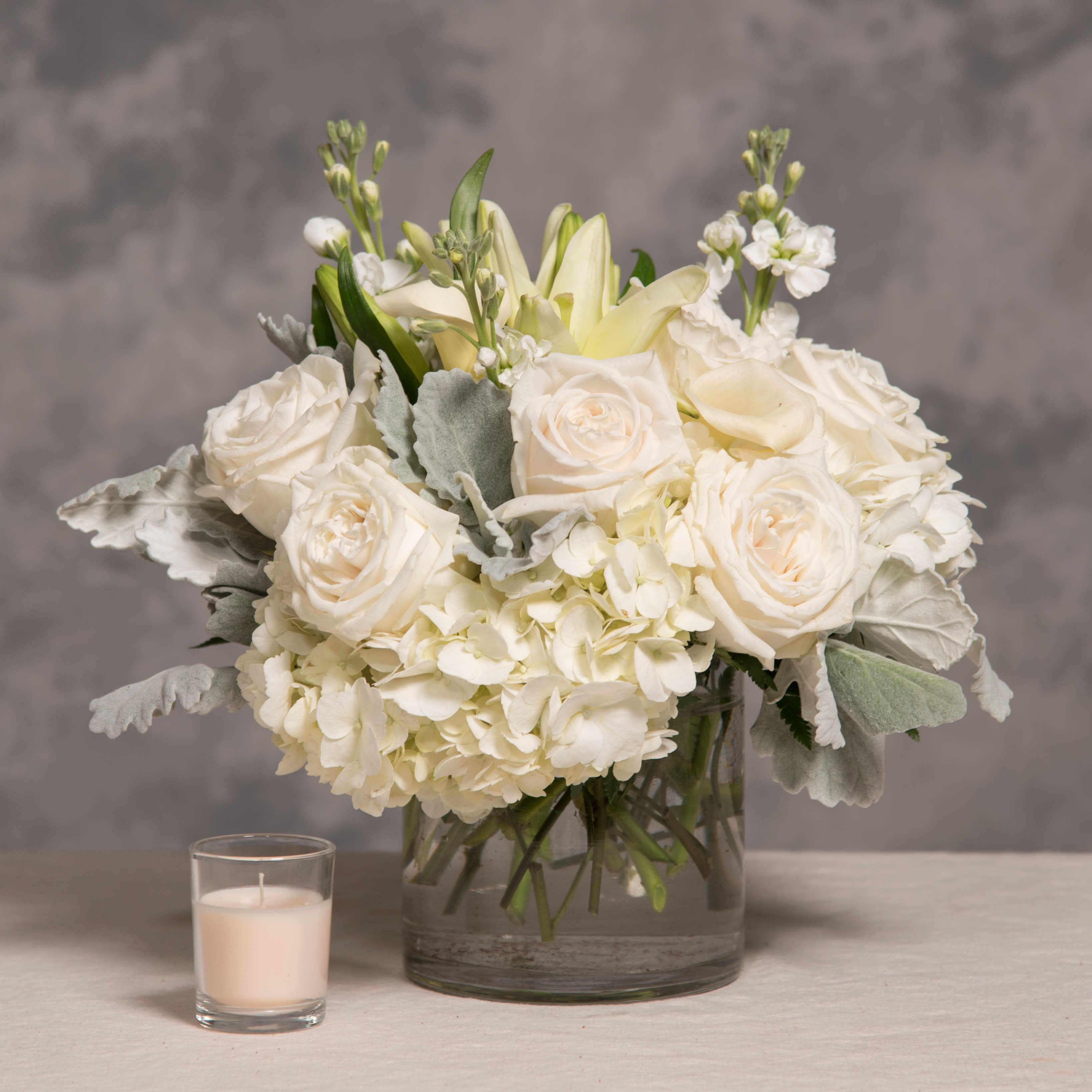 White roses and hydrangeas in a glass vase with a small candle beside it