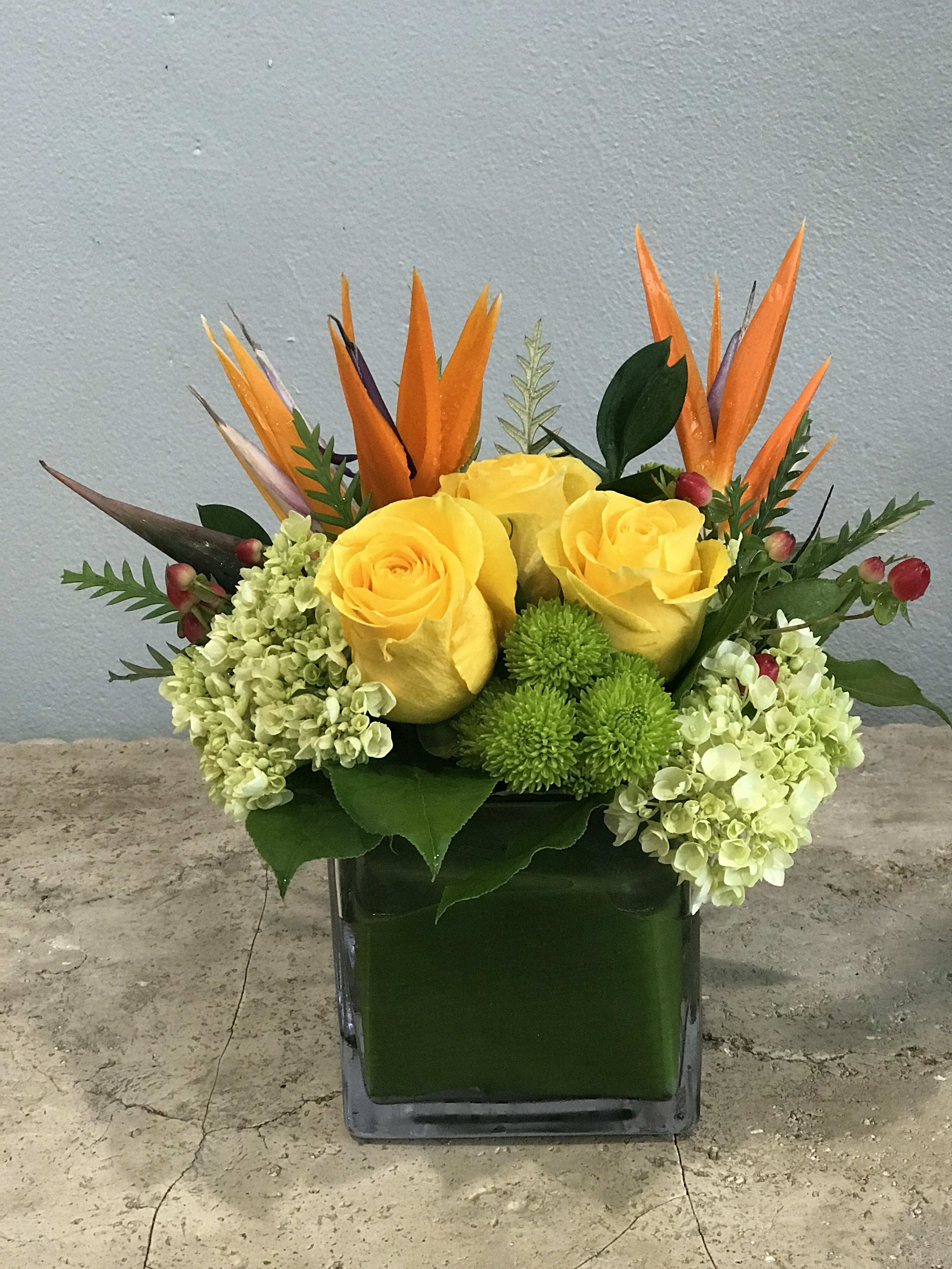 Yellow roses and green hydrangeas in a square glass vase with orange tropical accents
