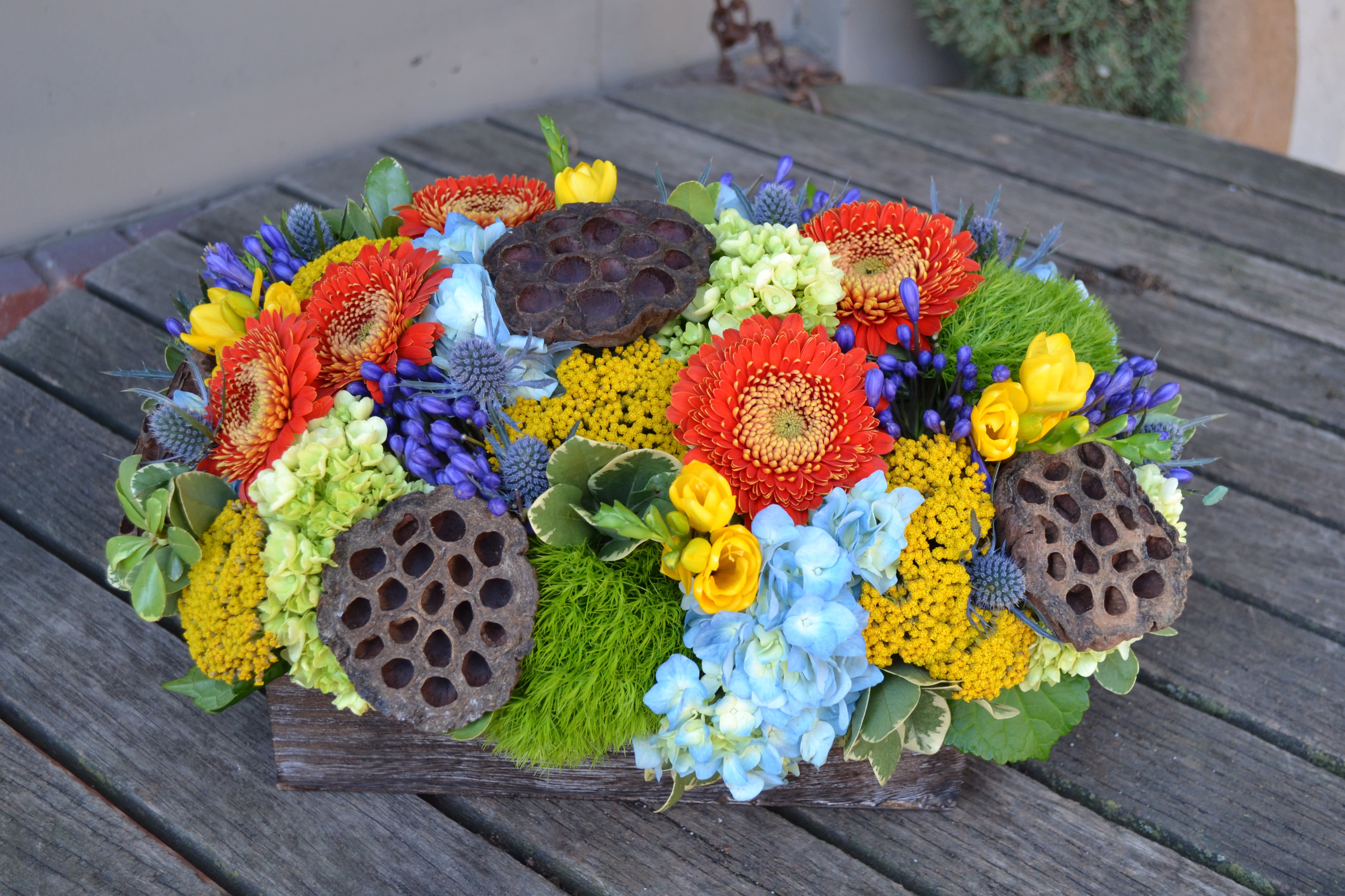 Low floral arrangement with red, yellow, blue, and green blooms in a wooden container