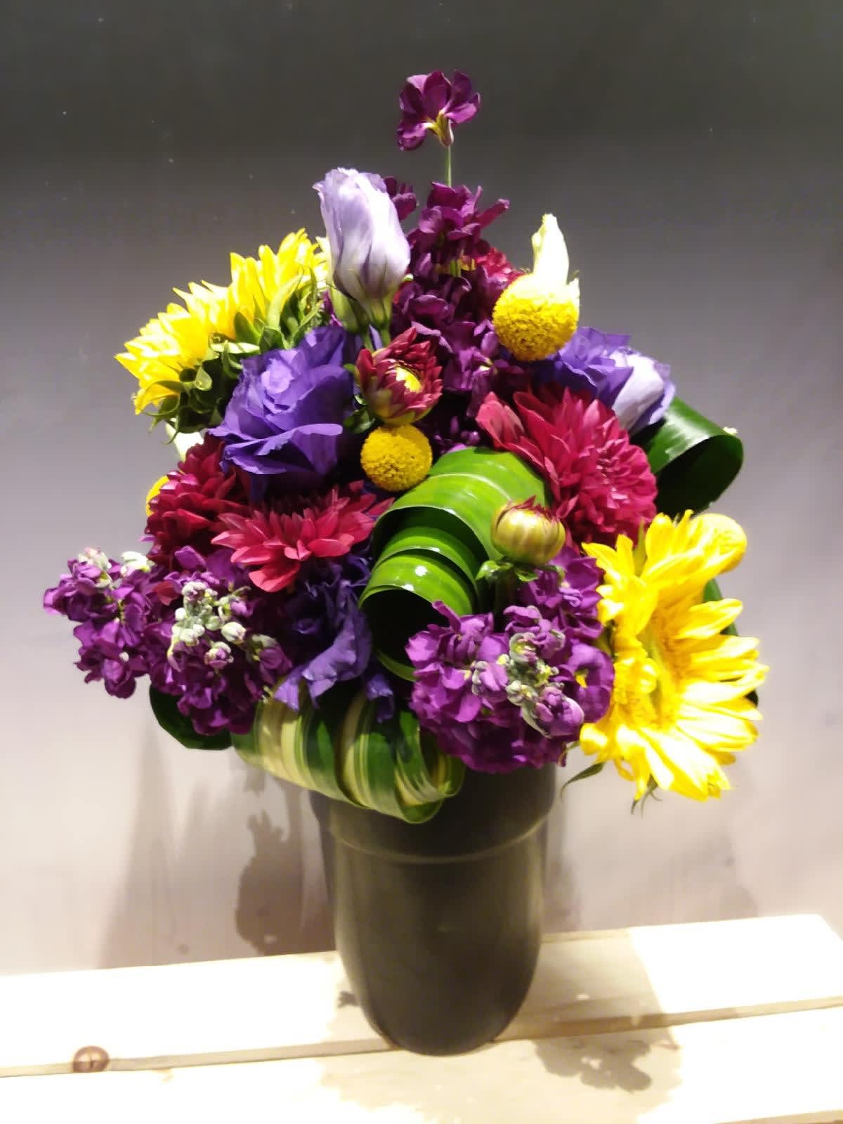 Tall mixed arrangement of yellow sunflowers, purple lisianthus, and magenta mums in a dark vase