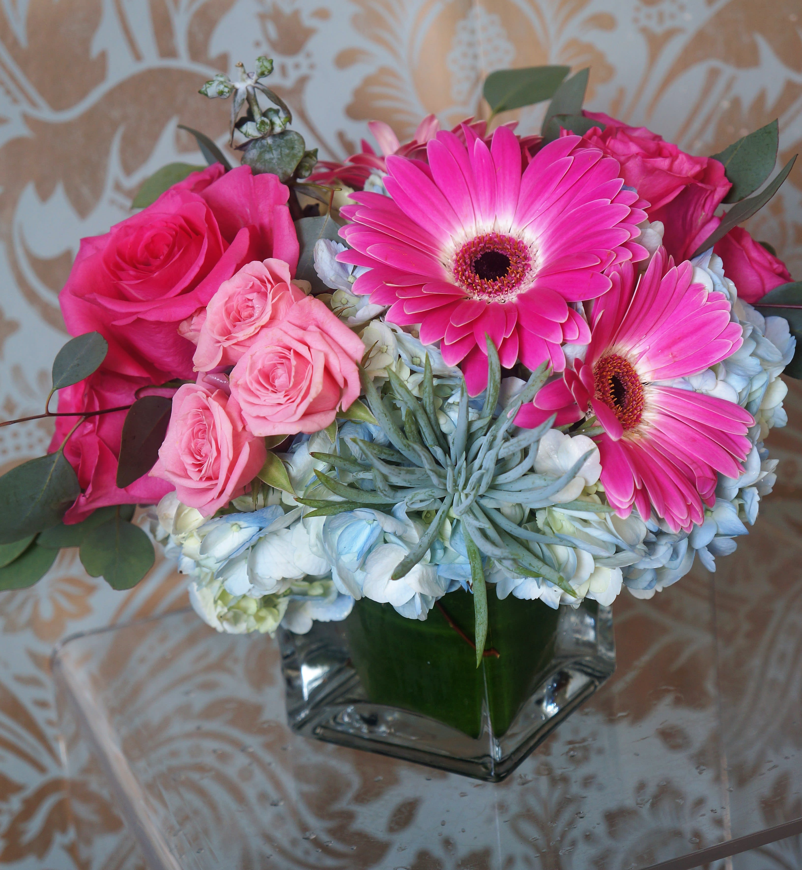 Low arrangement of bright pink gerbera daisies, roses, and hydrangeas in a square glass vase.