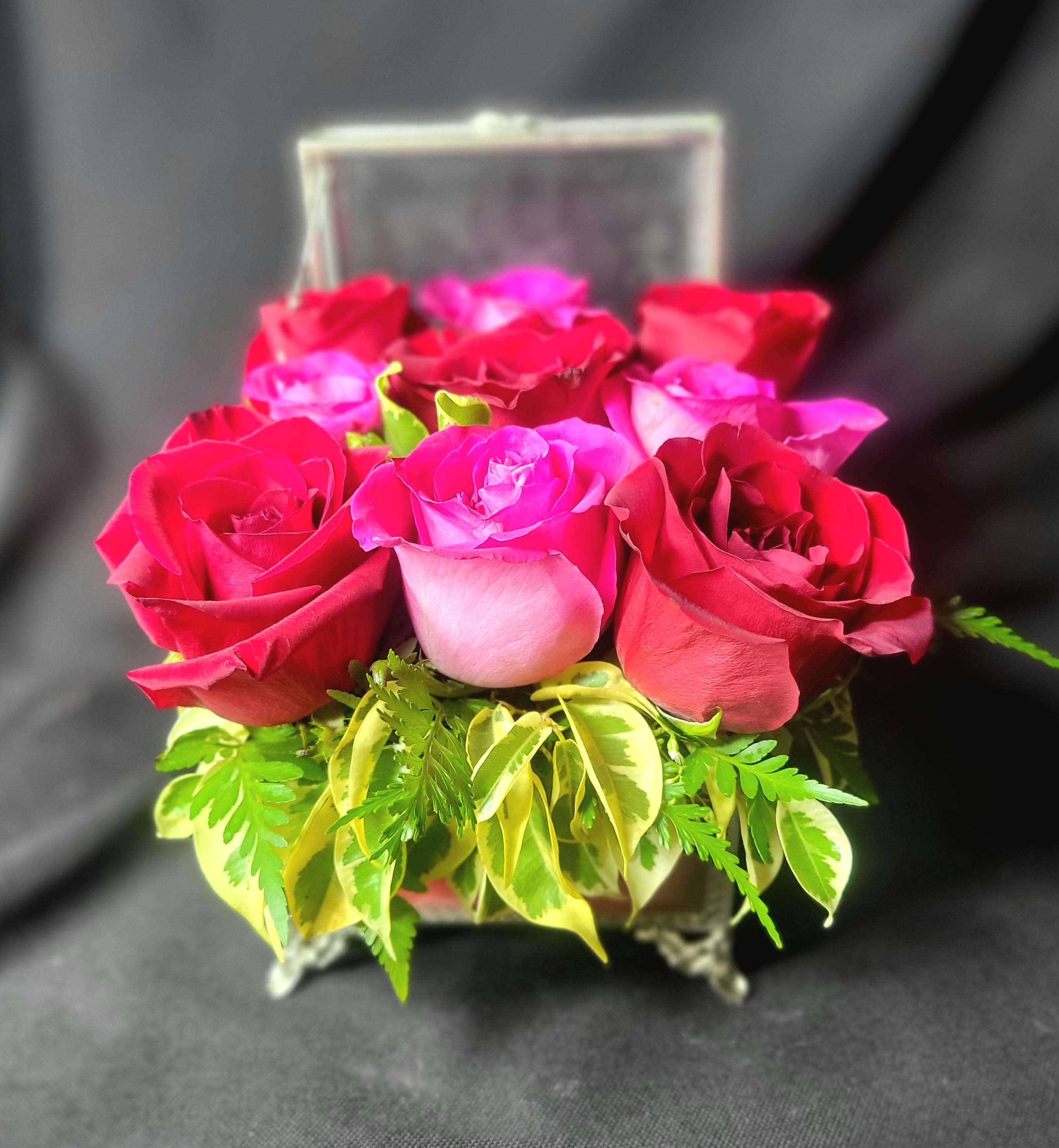 Compact arrangement of red and pink roses in a clear glass box with variegated foliage at the base