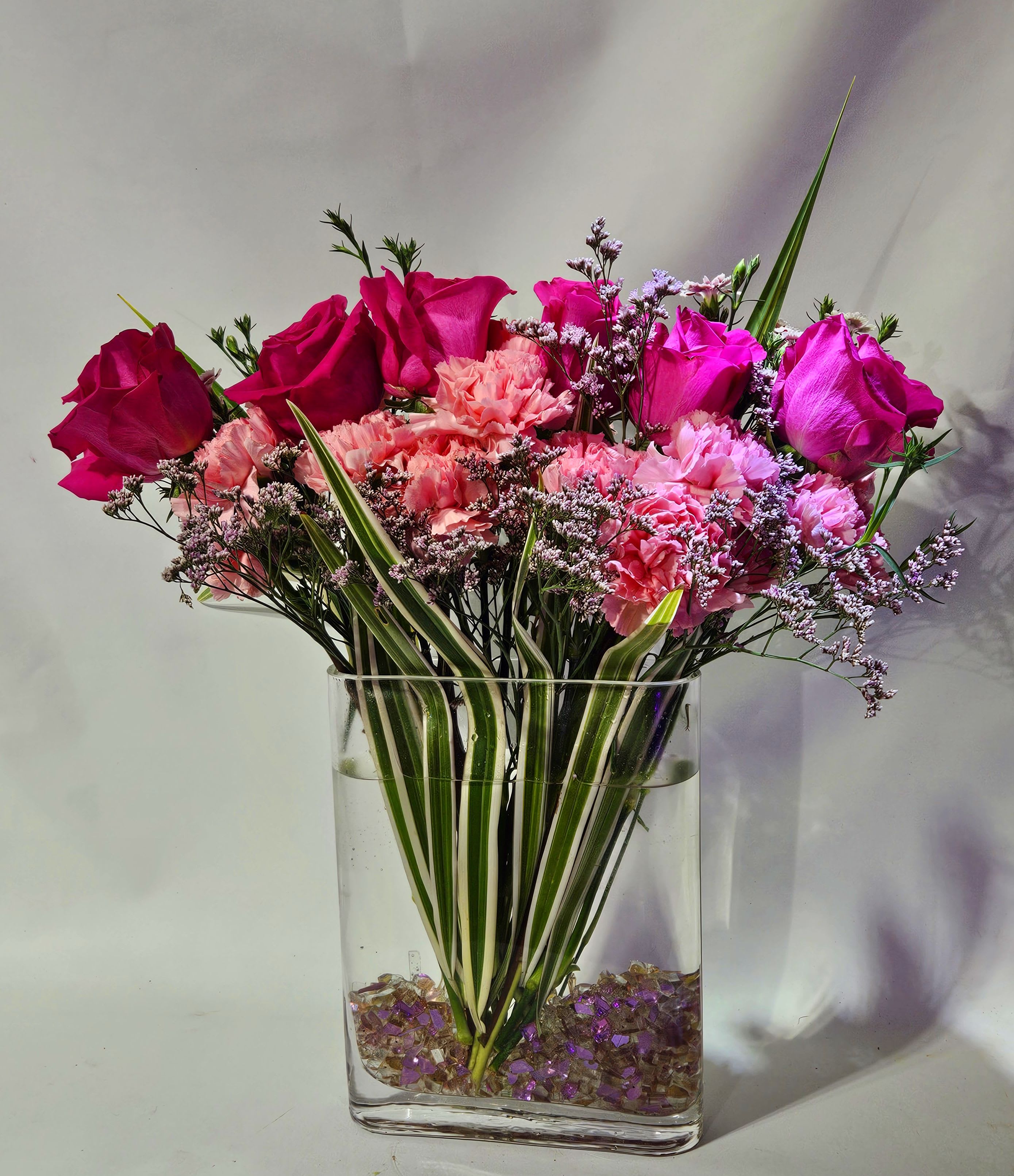 Tall arrangement of hot pink roses and pink carnations in a clear rectangular vase with striped foliage.
