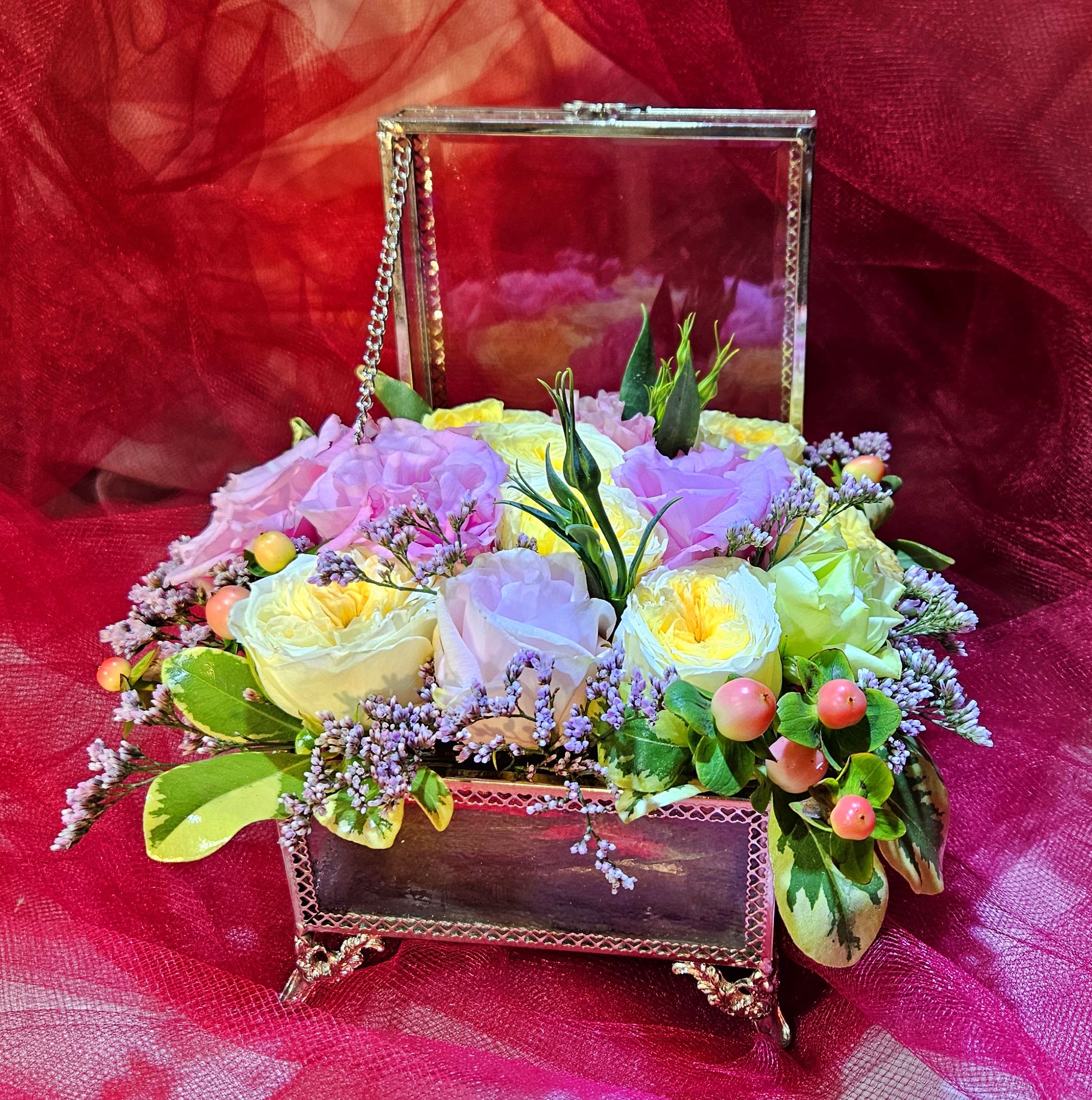 Pastel roses and filler flowers arranged in an open glass jewelry-style box on a red fabric backdrop.