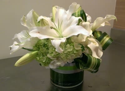 Low white lily and green hydrangea arrangement in a clear glass cylinder vase with looped striped foliage.
