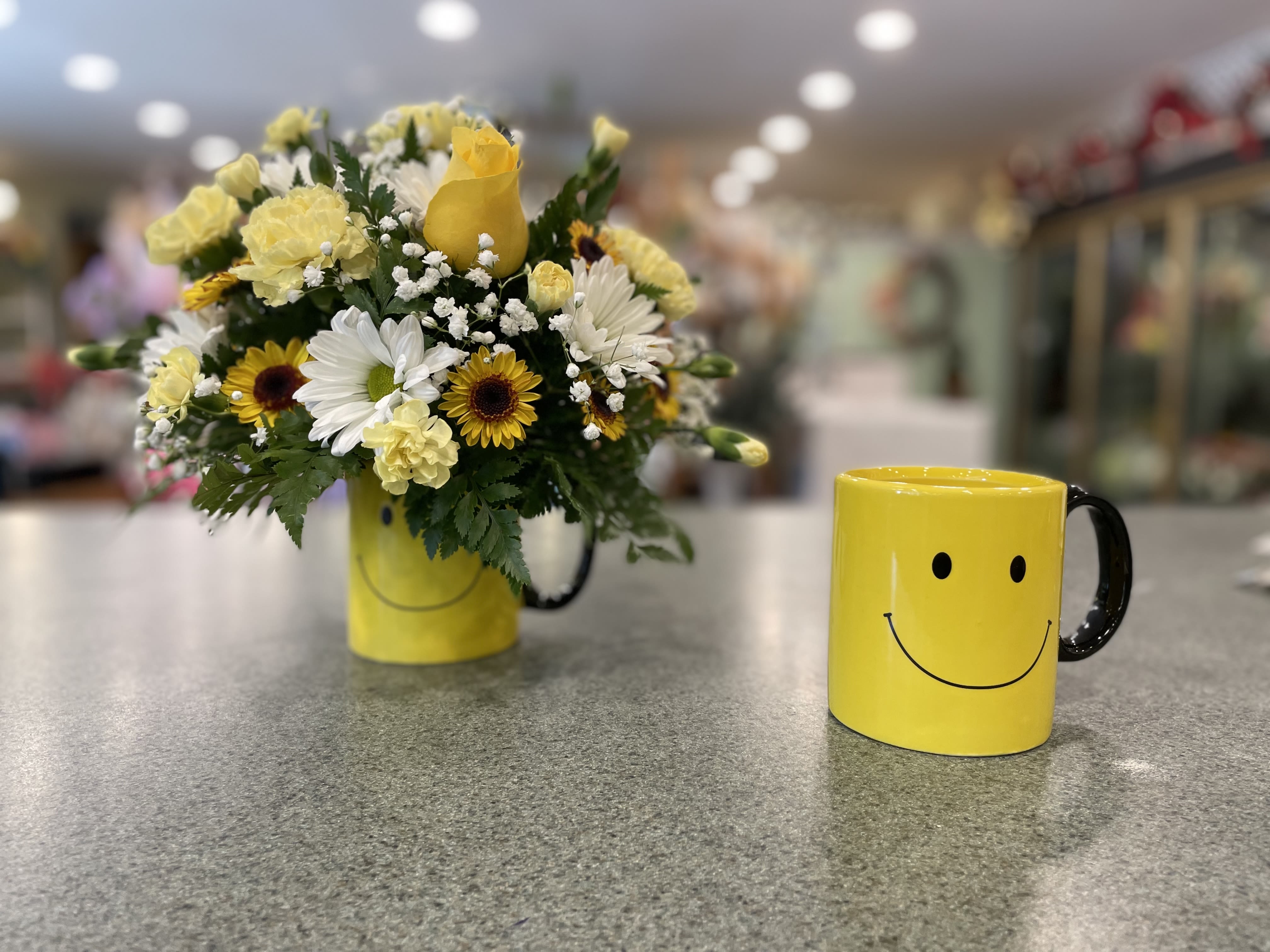 Yellow and white flower arrangement in a smiley-face mug beside a matching mug