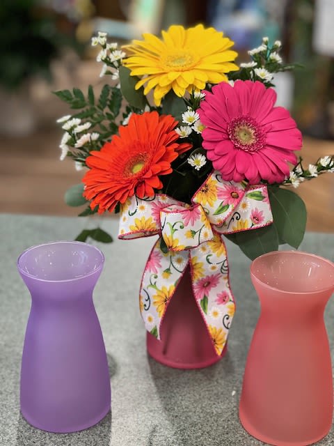 Bright gerbera daisies in a frosted pink vase with floral bow, flanked by two empty frosted vases.