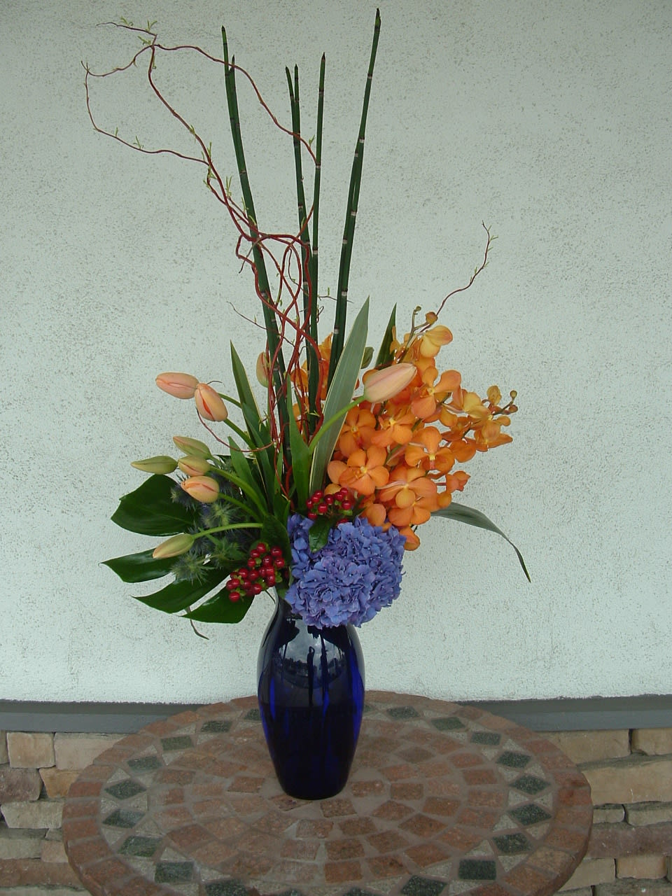 Tall floral arrangement in a blue vase with orange orchids and blue hydrangea