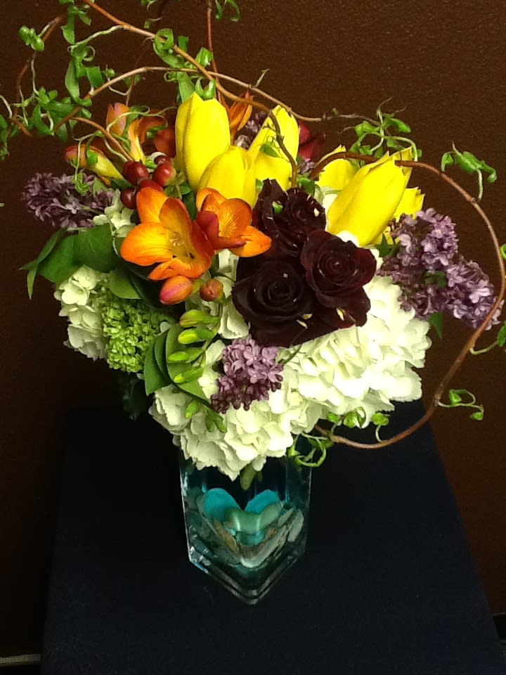 Colorful bouquet with yellow tulips, orange blooms, and dark roses in a glass vase