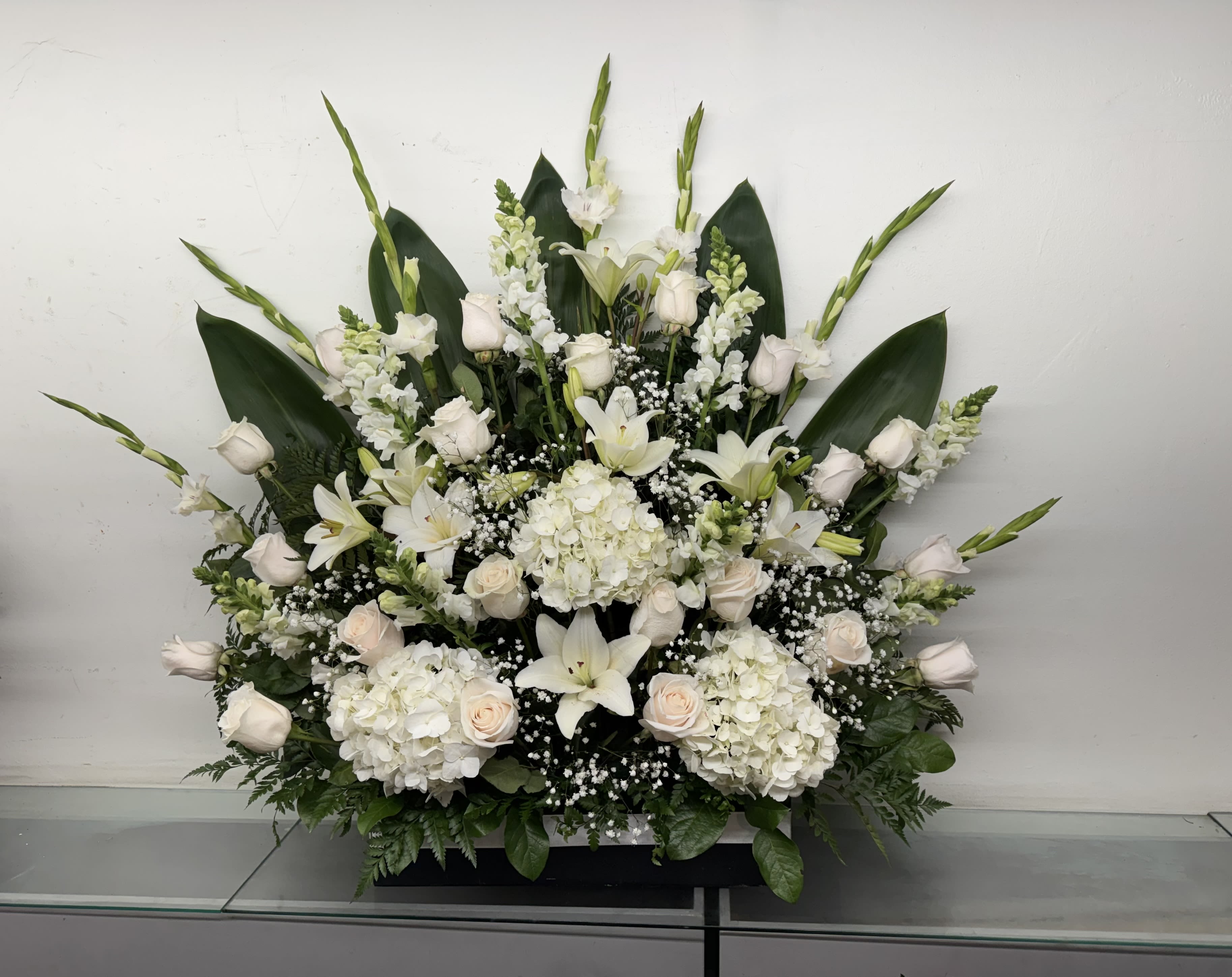 Ivory Grace - Pure Elegance is a stunning one-sided floral arrangement designed to make a sophisticated statement. Featuring luxurious white hydrangeas, creamy white roses, lilies, snapdragons, baby's breath, and lush greenery, this arrangement showcases timeless beauty and refined style.  Its impressive height and elegant silhouette make it an excellent choice for podium displays, church services, corporate events, anniversaries, celebrations of life, and meaningful occasions where a tasteful floral presentation is desired. The soft white palette symbolizes grace, peace, sincerity, and admiration, creating a display that is both striking and versatile.