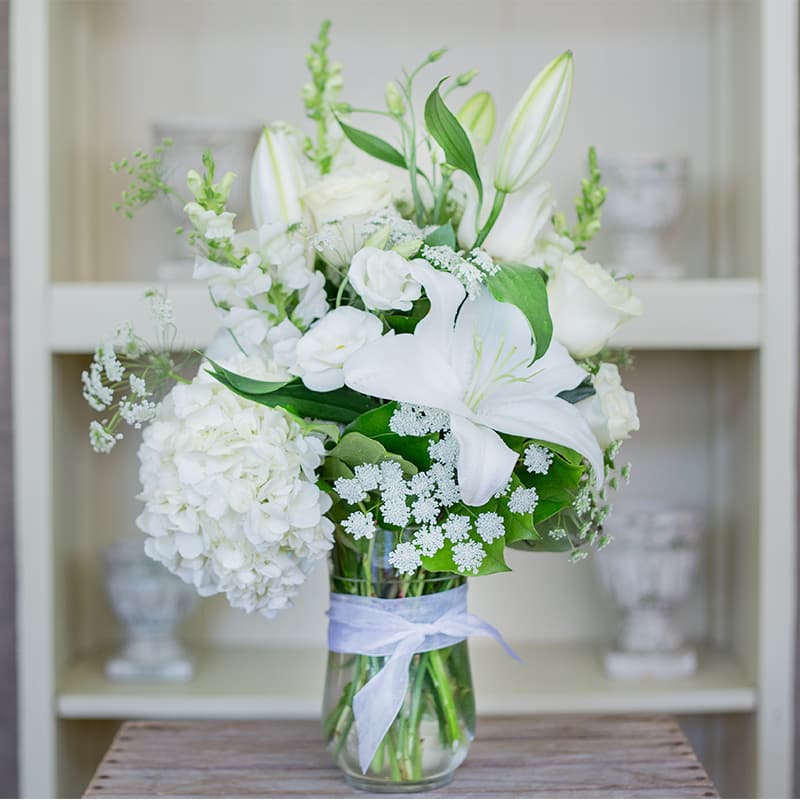 White lilies, roses, and hydrangea arranged in a clear glass vase tied with a white ribbon