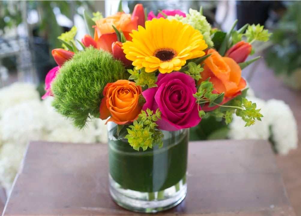 Bright bouquet of yellow gerbera, orange and pink roses, and green blooms in a clear glass vase
