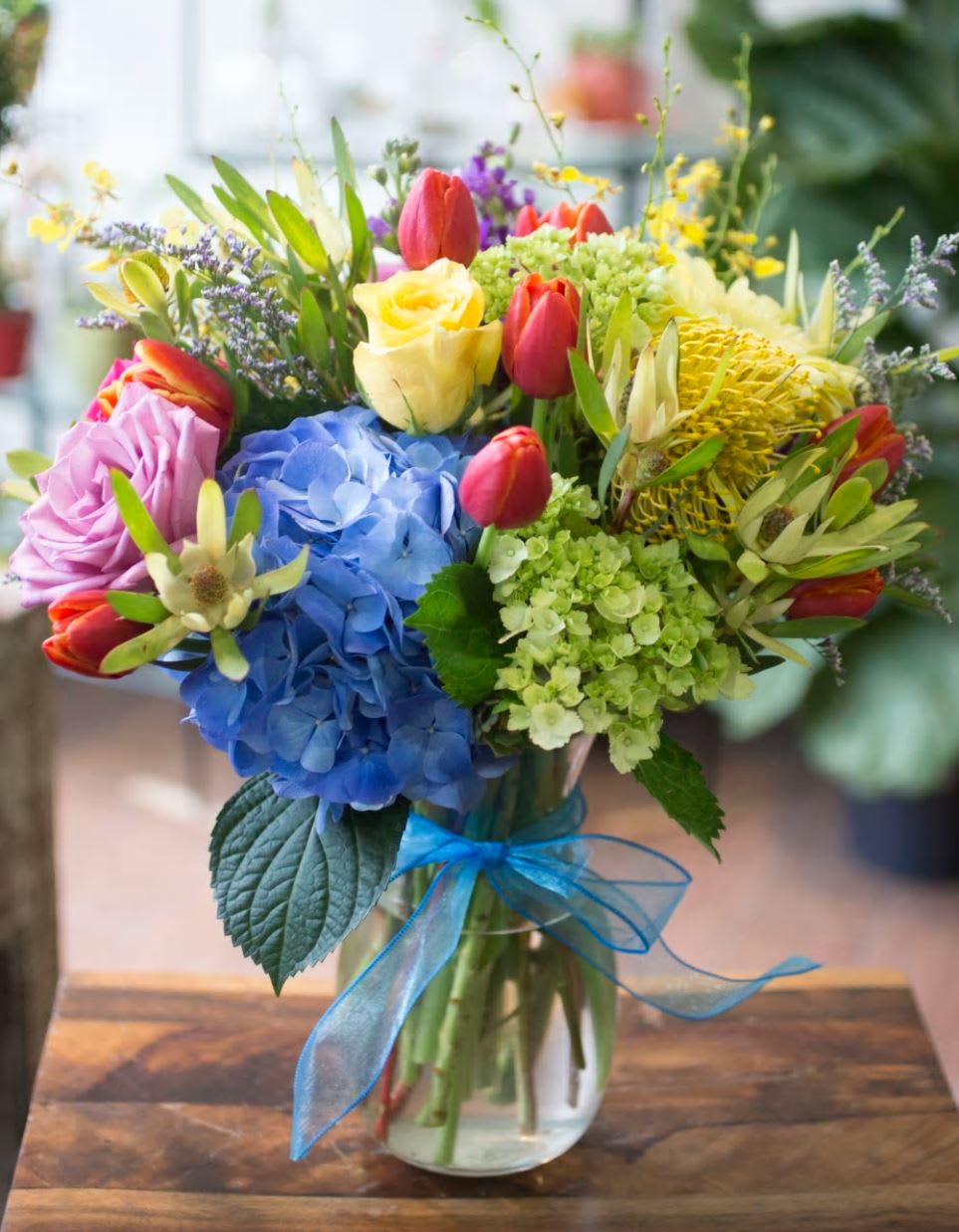 Colorful bouquet with blue hydrangea, yellow rose, red tulips in a glass vase with blue ribbon