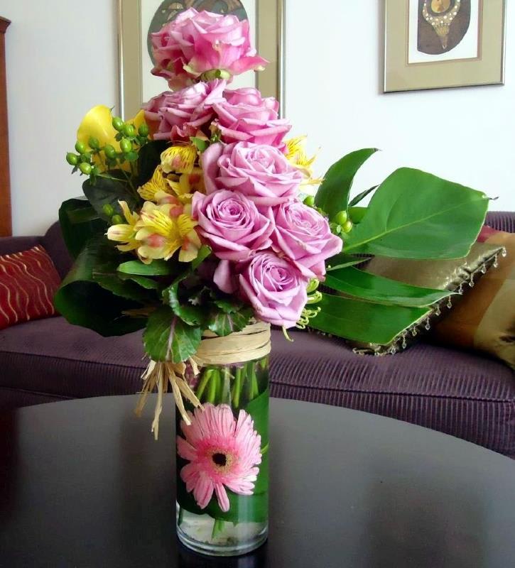 Tall glass vase with pink roses, yellow blooms, and a pink gerbera daisy framed by bold green leaves.