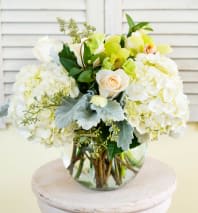 Round arrangement of white hydrangeas, pale roses, and green orchids in a clear glass vase