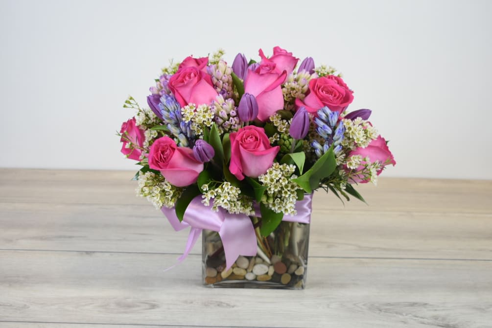 Compact arrangement of pink roses and purple flowers in a square glass vase with stones and a satin ribbon.
