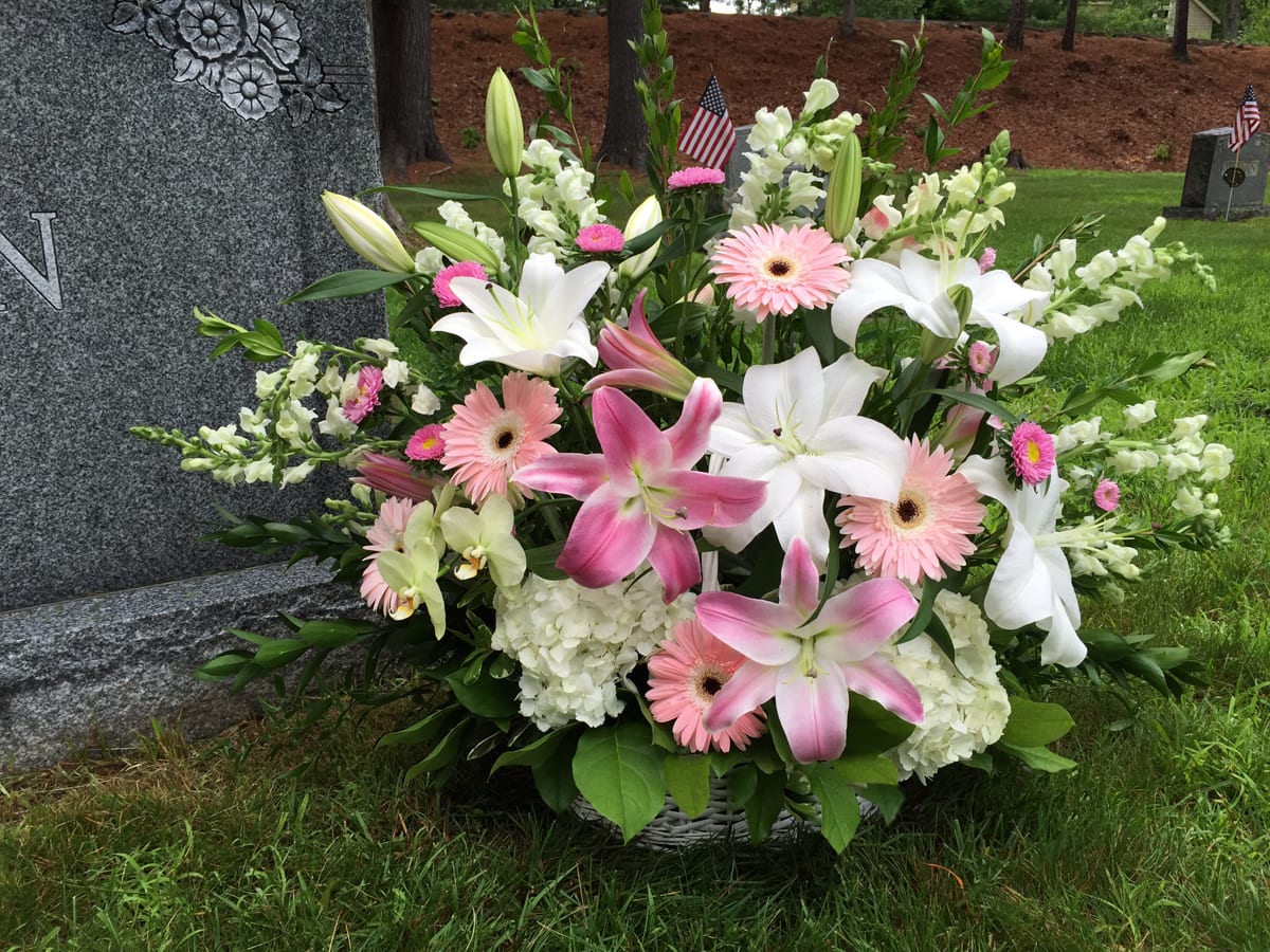 Pink and white lily and gerbera daisy basket arrangement set beside a headstone.