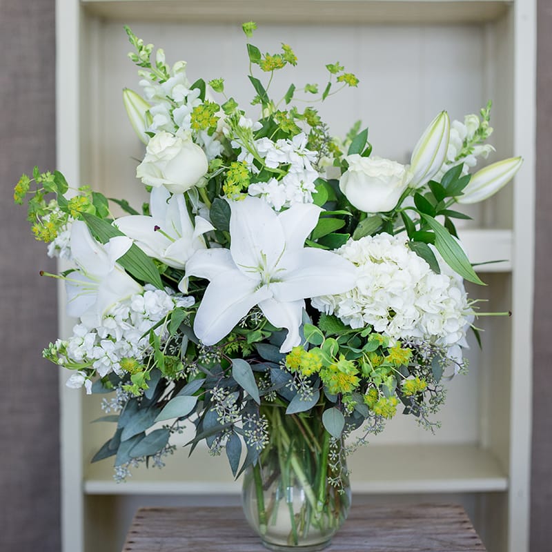 Tall arrangement of white lilies, roses, hydrangeas, and yellow-green blooms in a clear glass vase