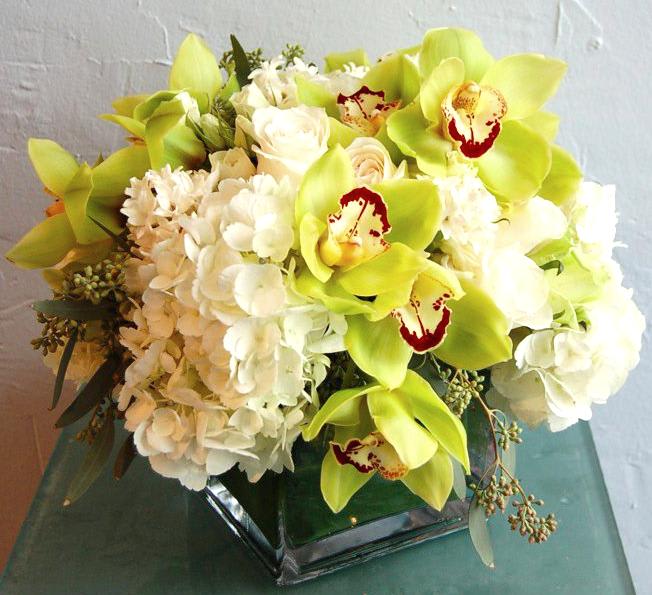 Low arrangement of green cymbidium orchids, white hydrangeas, and roses in a clear glass cube vase