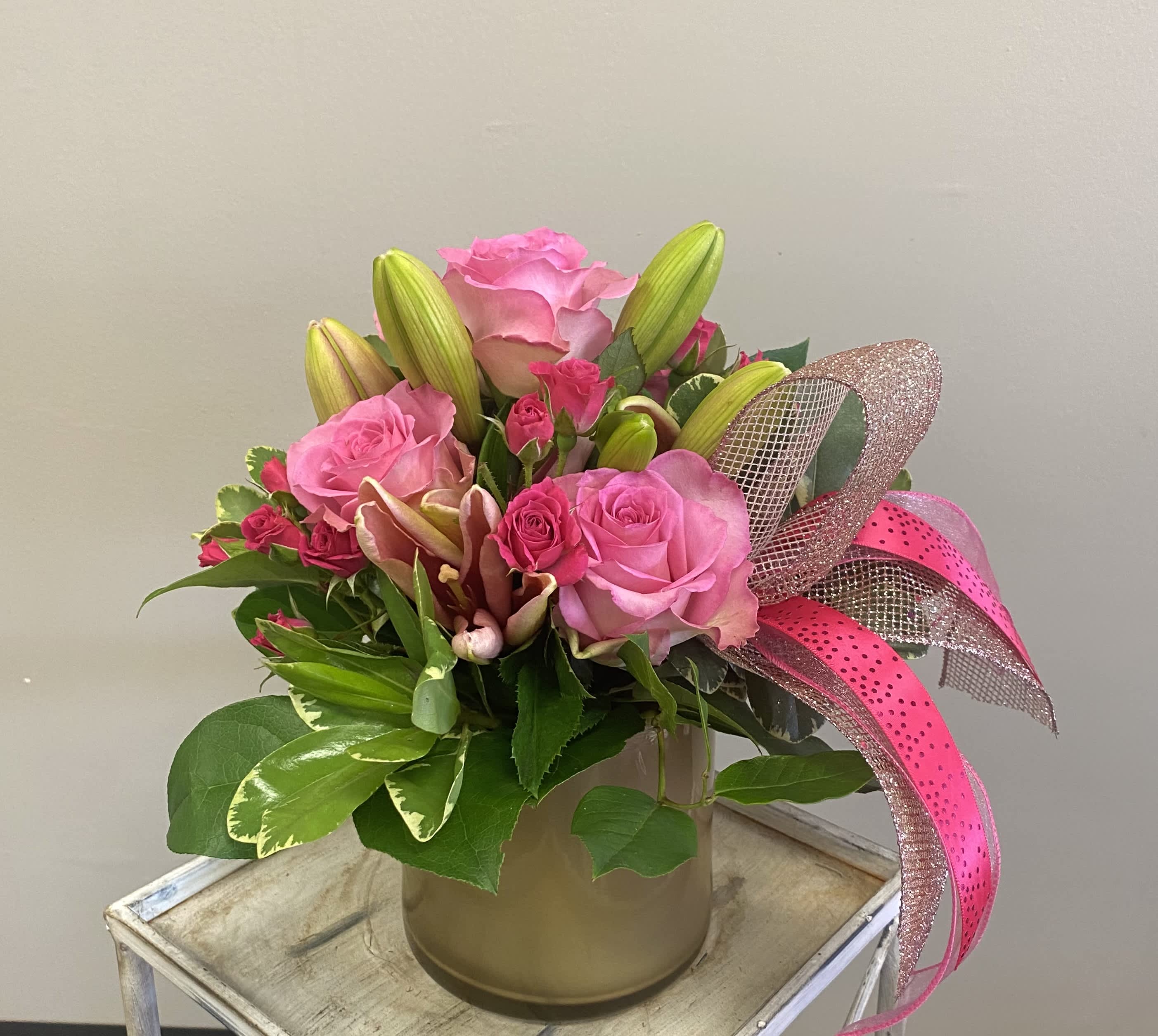 Pink roses and lilies arranged in a gold vase with a pink ribbon