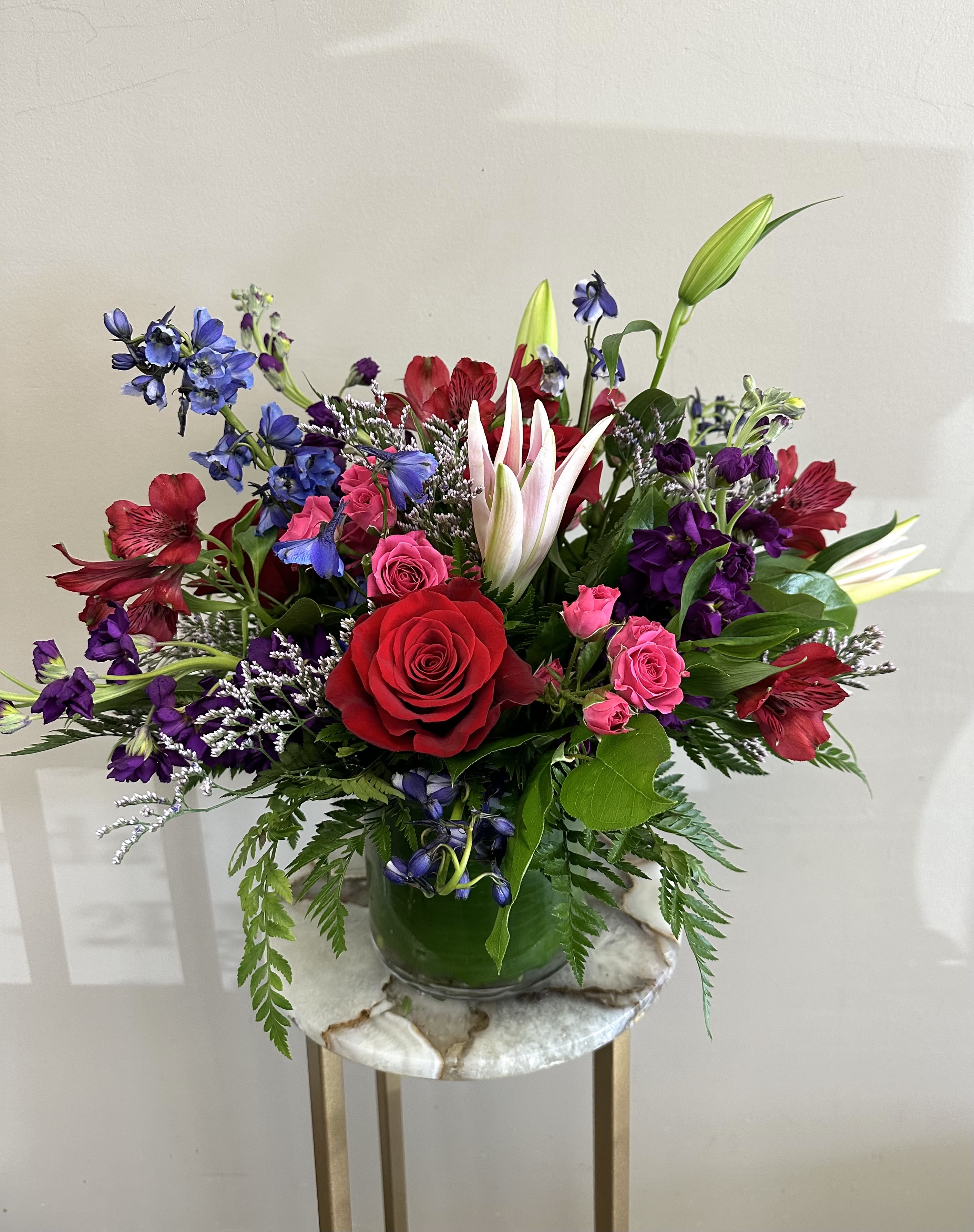 Colorful mixed arrangement with red roses, pink lilies, and purple blooms in a clear glass vase