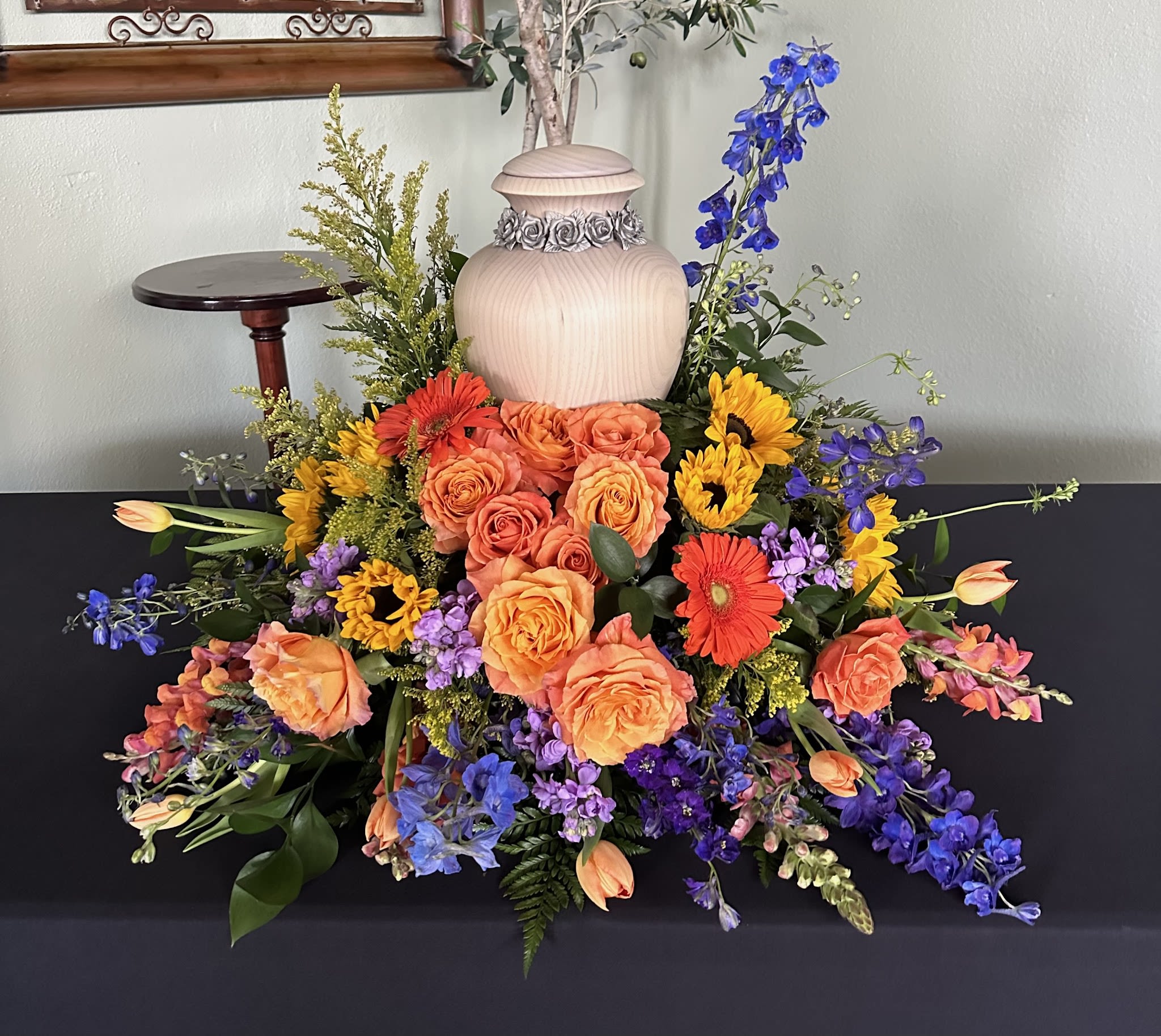 Colorful floral arrangement with roses, gerbera daisies, and blue delphinium around an urn