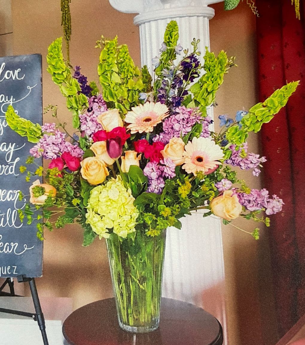 Tall mixed arrangement of pink, peach, and green flowers in a clear glass vase on a table