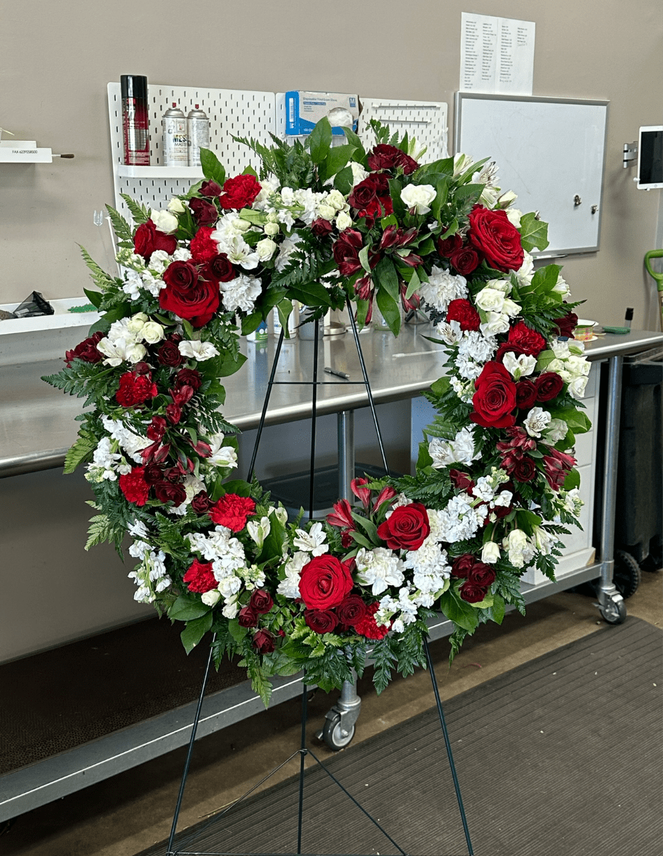 Wreath WR-07 - A timeless expression of love and remembrance, this standing wreath features a rich mix of red roses, deep burgundy blooms, and soft white carnations, accented with lush greenery. Its classic color palette makes it a dignified tribute for memorial services in Estrella Mountain Ranch, Verrado, and surrounding West Valley communities. Easel stand included; ribbon lettering available for an additional charge.