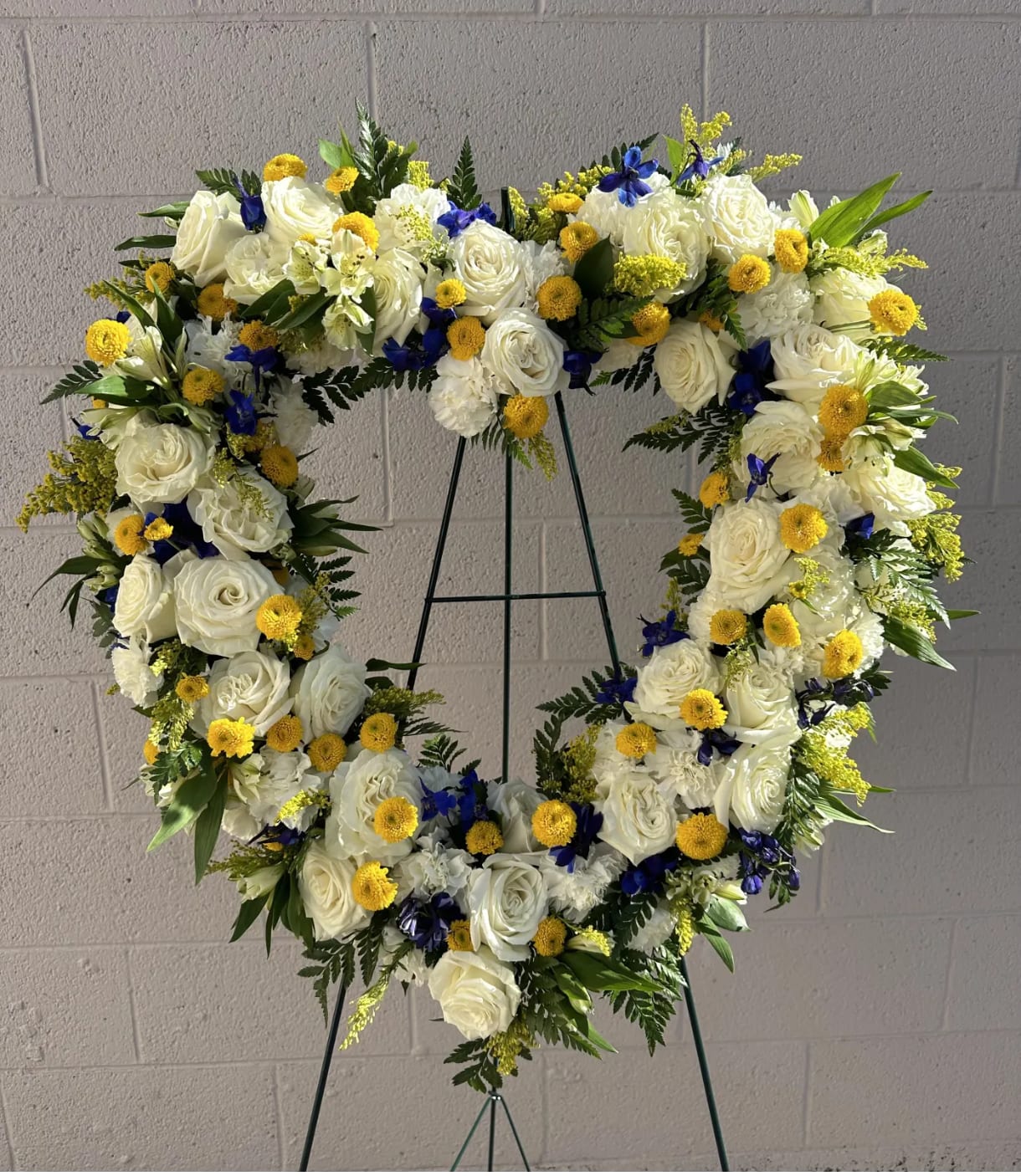 Heart-shaped floral wreath with white roses, yellow blooms, and blue accents on a stand