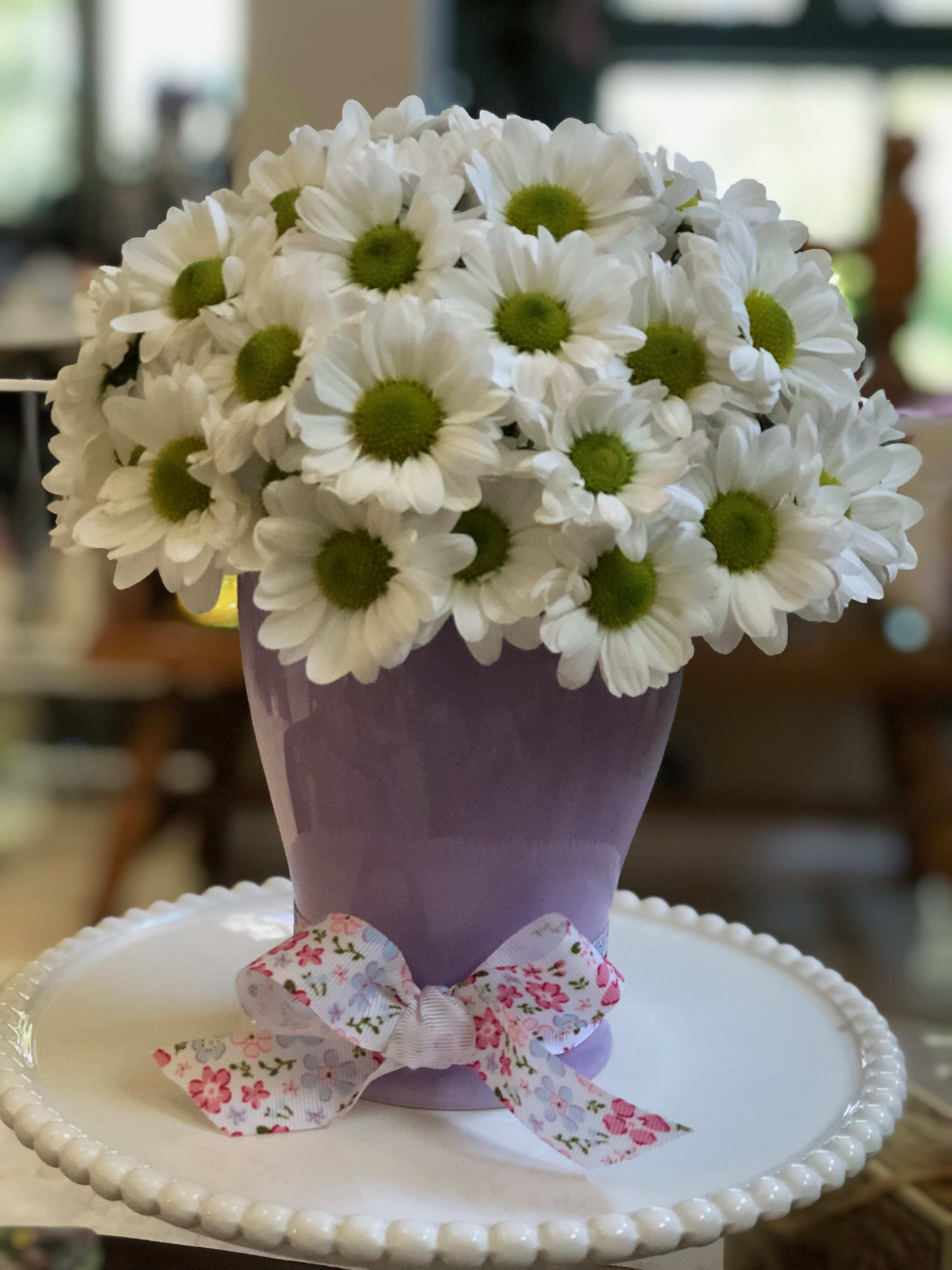 White daisies in a lavender vase with a floral ribbon bow