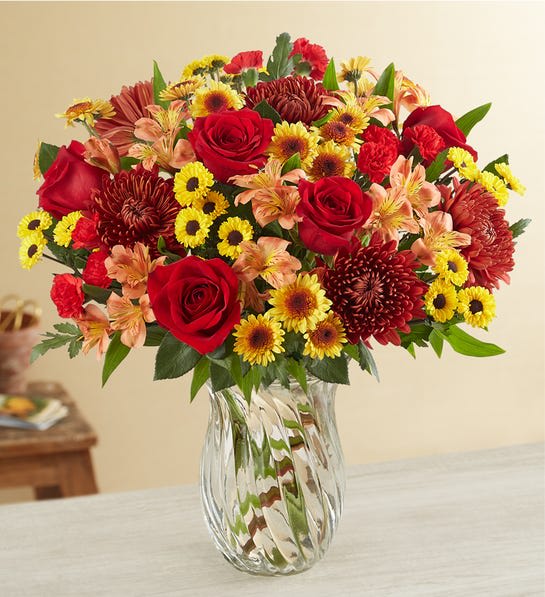 Mixed bouquet of red roses, yellow daisies, and orange flowers in a clear glass vase
