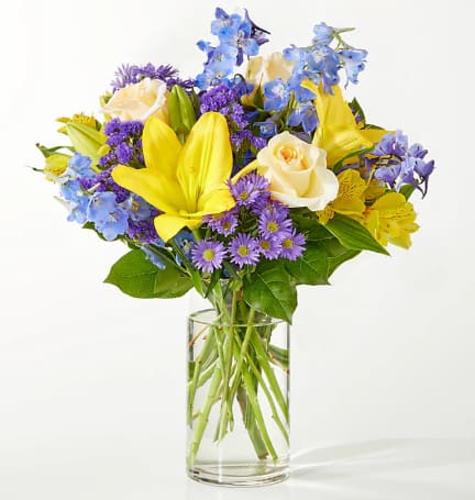 Bouquet of yellow lilies, cream roses, and purple flowers in a clear glass vase