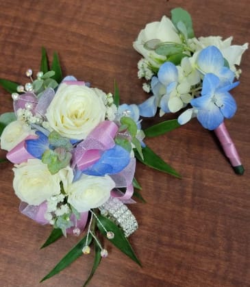 Spring Bling Corsage - Four white spray roses with a hint of light blue flowers and babies breath wrapped in pink ribbon and diamond like wristlet give this a spring w/ bling feel. The deluxe version includes a matching boutonniere. The roses and flowers can come in a variety of colors, include color choice in the notes if ordering online. it also can come in fresh or silk flowers.