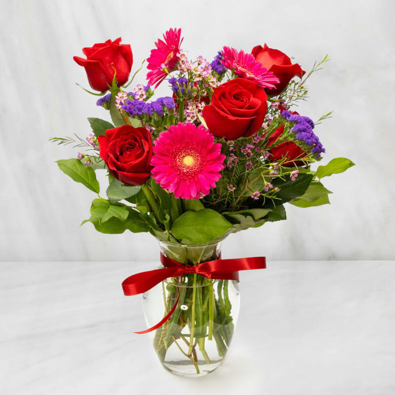 Red roses and pink gerbera daisies in a glass vase with a red ribbon