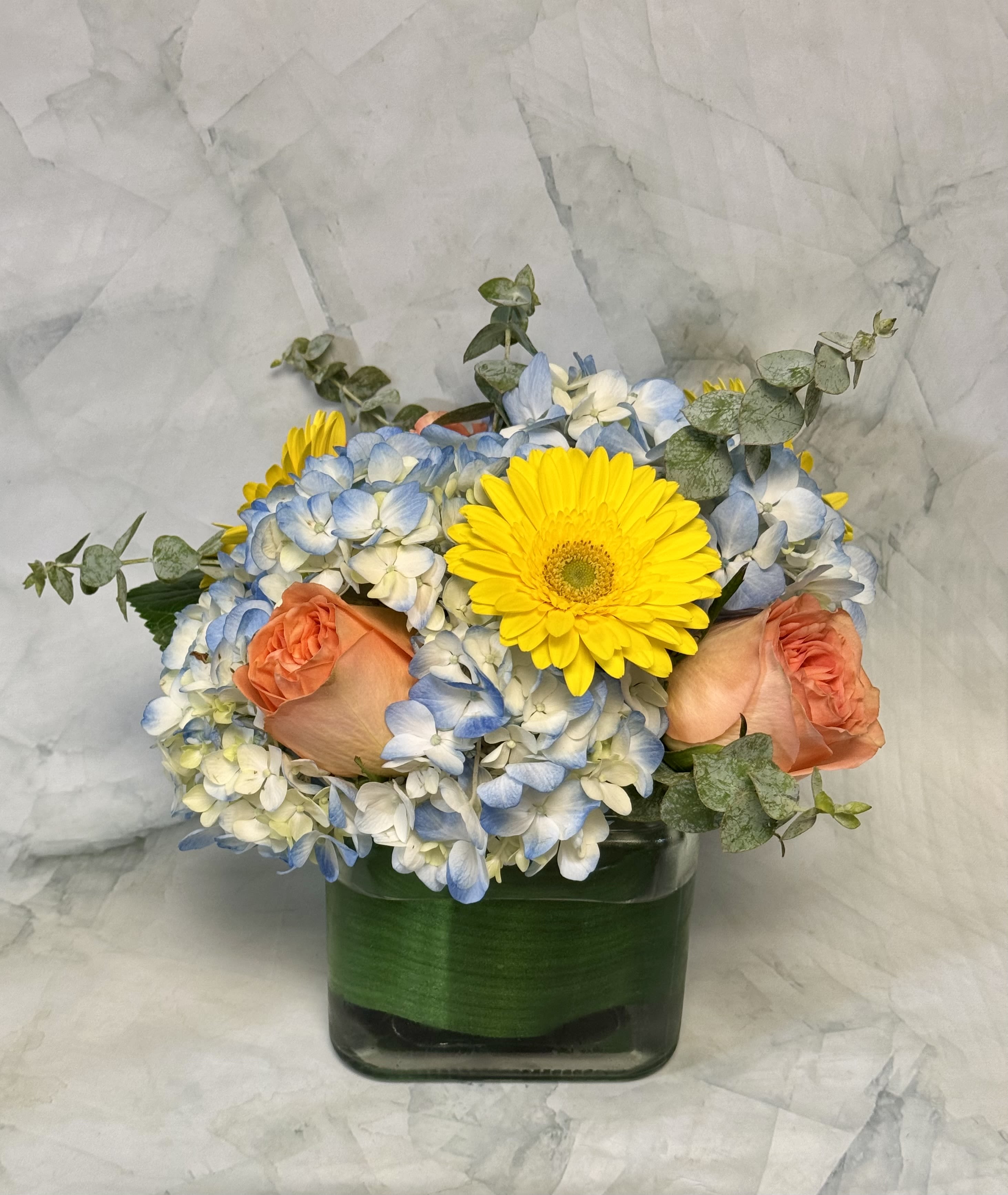 Bouquet of blue and white hydrangeas with yellow gerbera daisies and peach roses in a glass vase