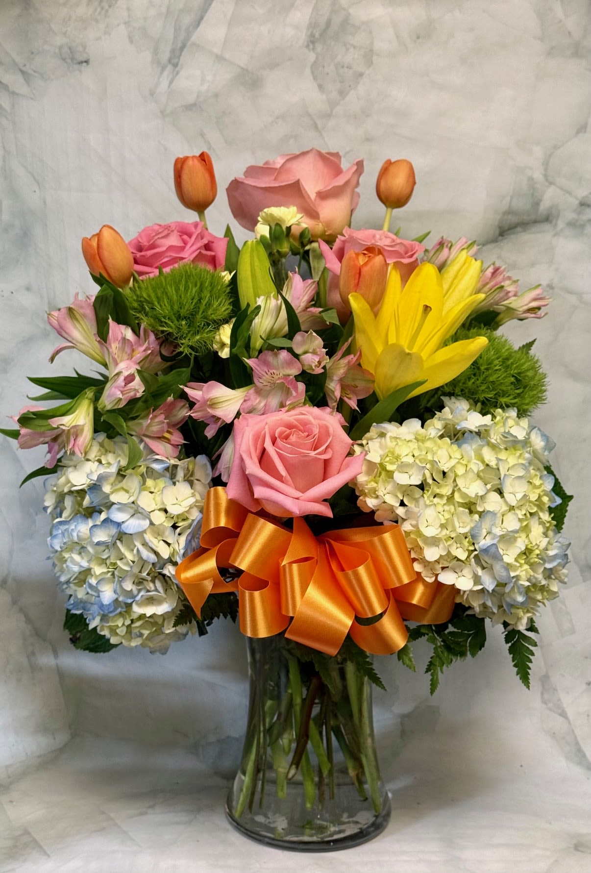 Tall mixed bouquet of pink roses, yellow lilies, tulips and hydrangeas in a clear vase with an orange bow.
