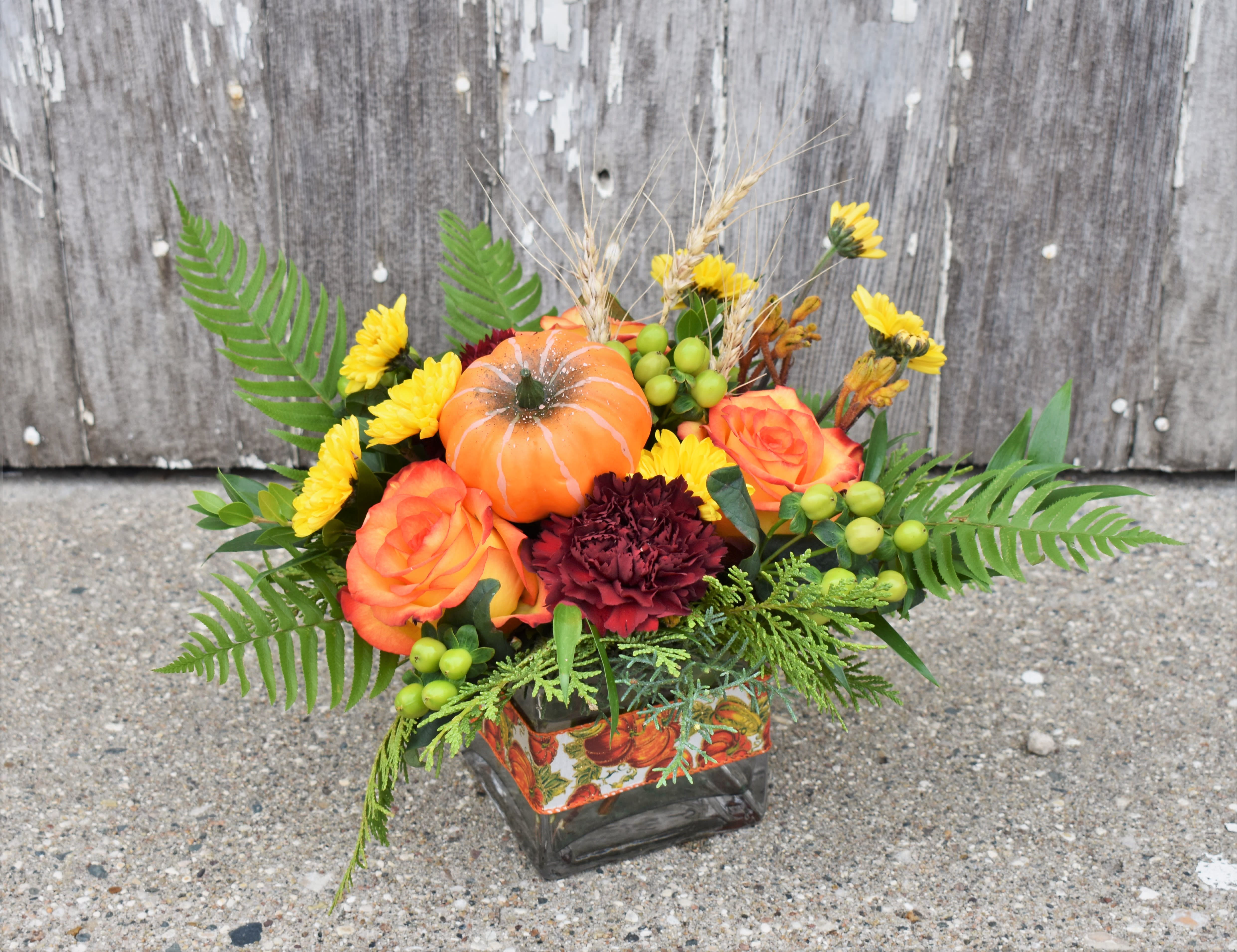 Autumn floral arrangement with orange roses, yellow daisies, and a small pumpkin in a square vase