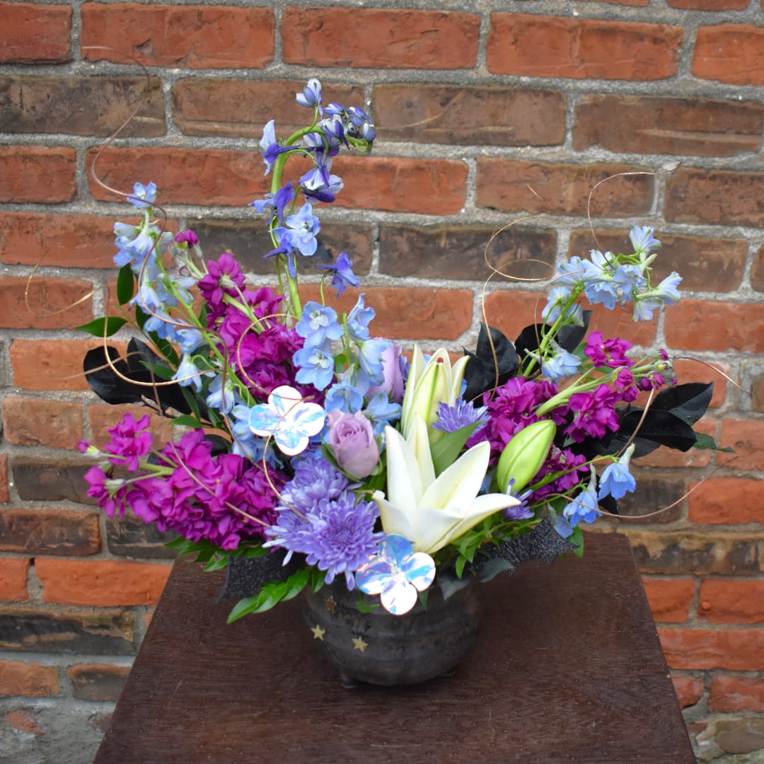 Mixed bouquet with white lilies, purple blooms, and blue flowers in a dark vase