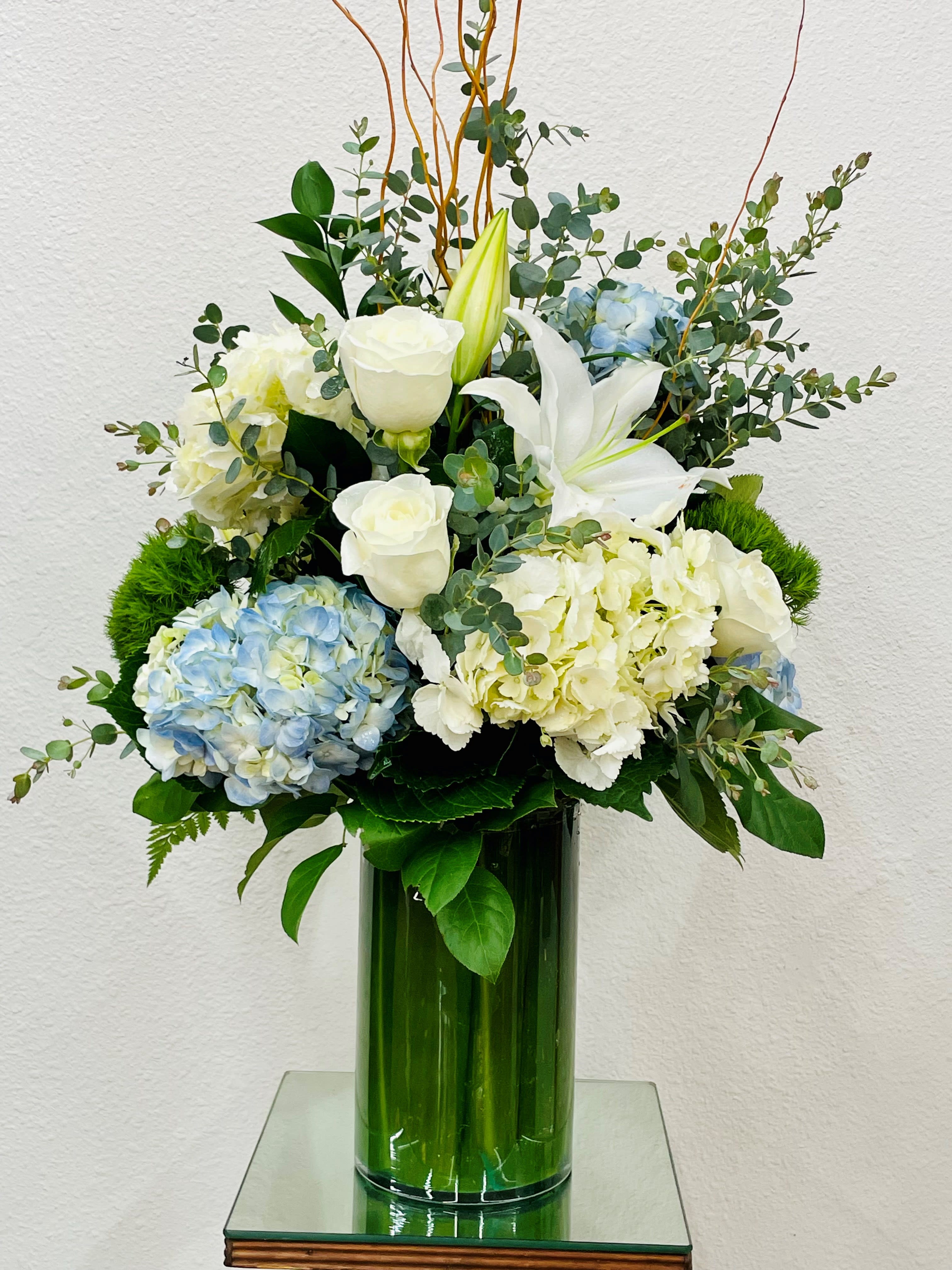 White lilies, roses, and blue hydrangeas in a tall green vase