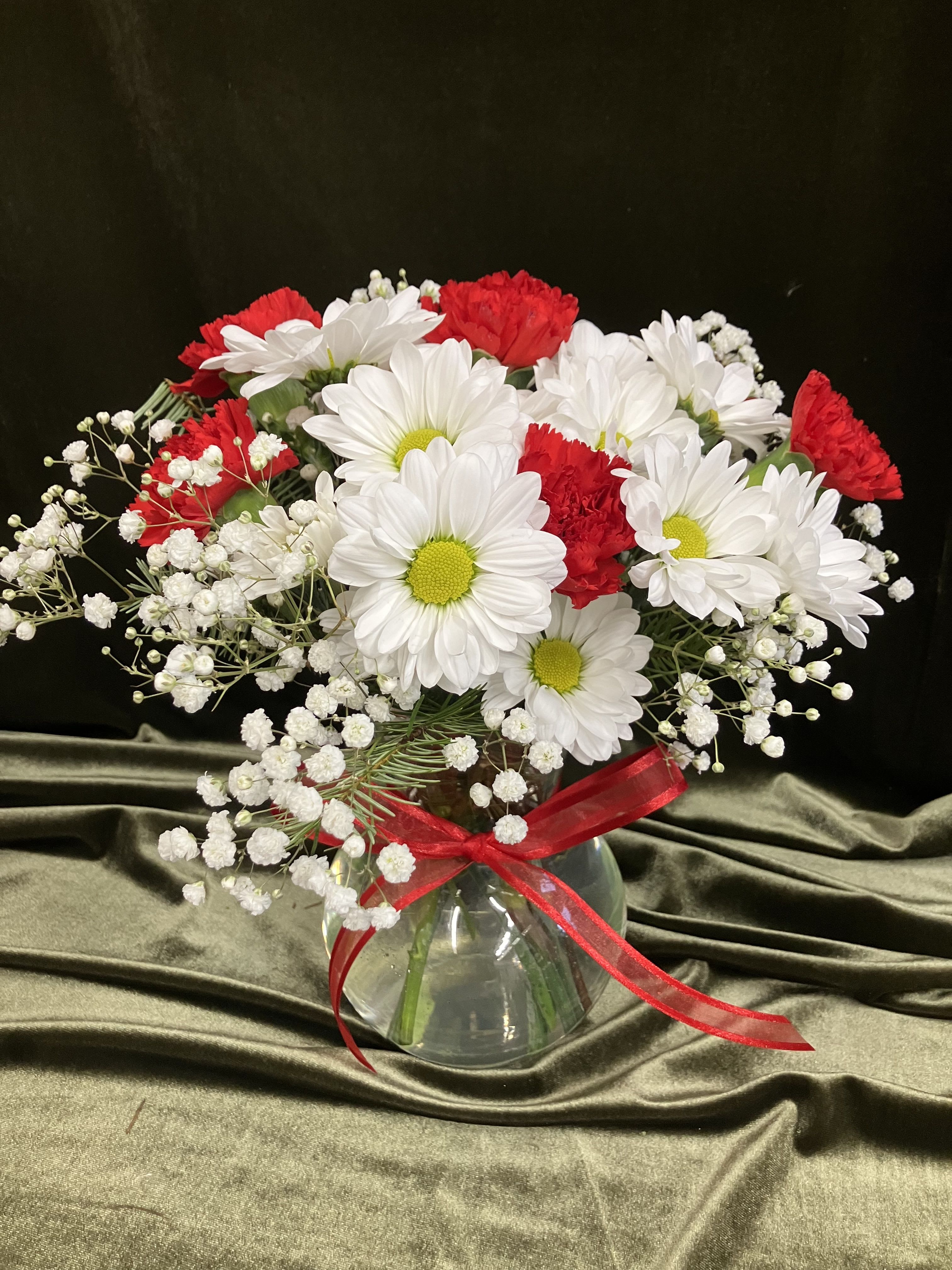 Red carnations and white daisies with baby's breath in a round glass vase with red ribbon